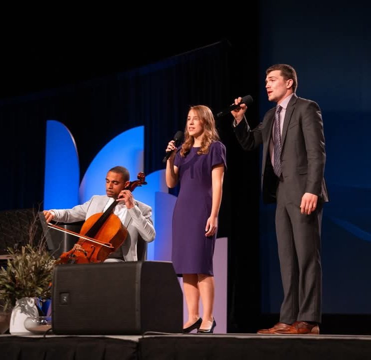 Three people performing on stage; a man playing the cello, a woman singing into a microphone, and a man singing into another microphone, with a blue backdrop.
