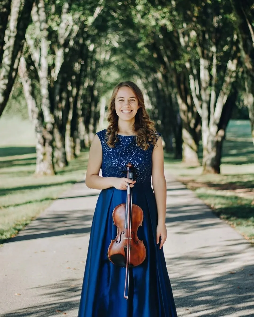 A young woman in a blue lace top and long blue skirt standing outdoors on a tree-lined sidewalk, smiling and holding a violin with a bow.