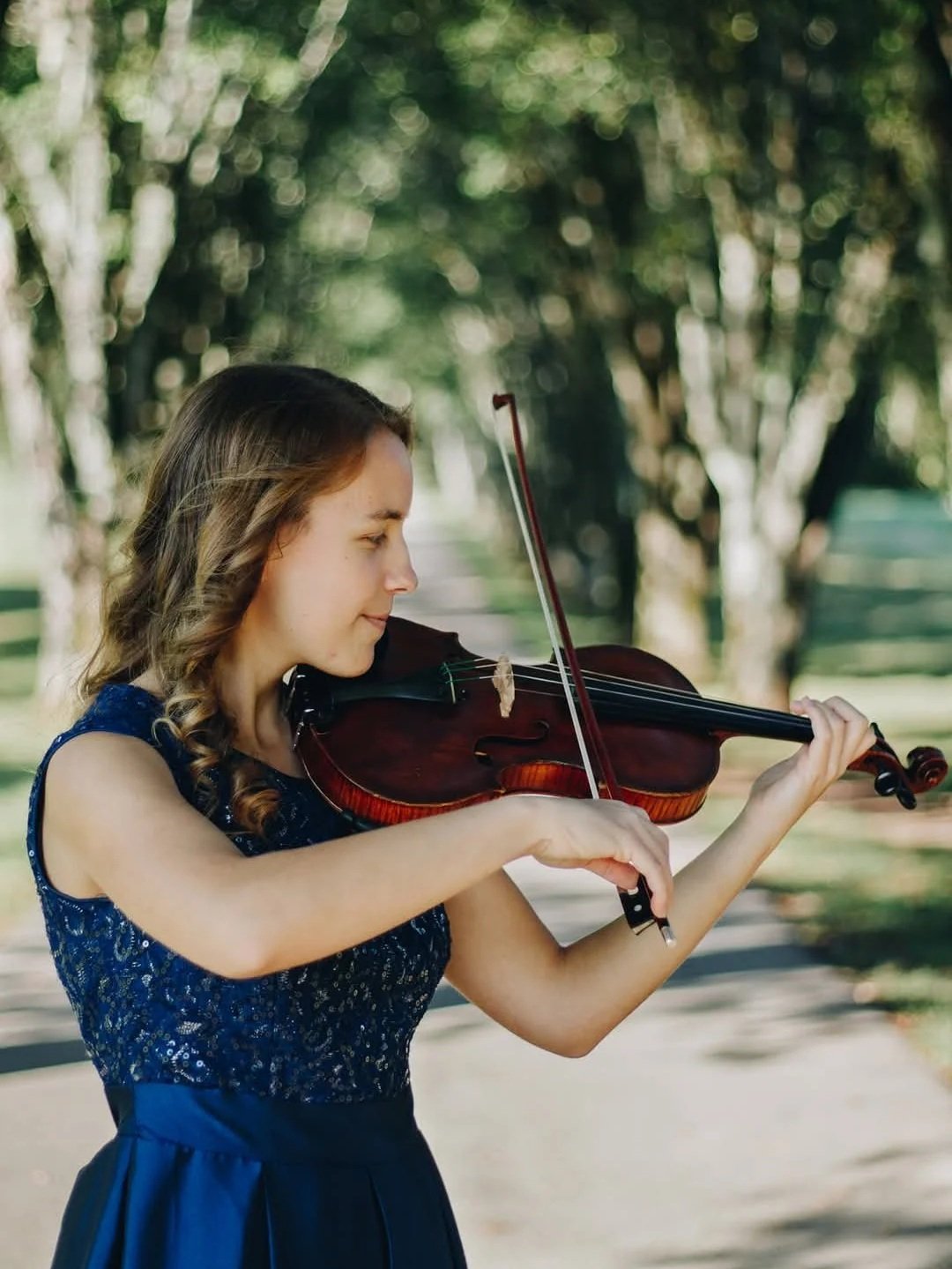 Young woman playing the violin outdoors in a park with trees in the background.