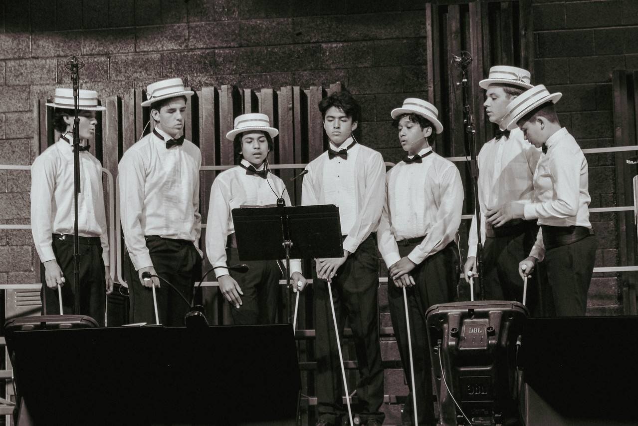 A group of seven young boys dressed in white shirts, black pants, and bow ties, wearing boater hats, standing on a stage with music stands and microphones, appearing to be in a choir or singing group.