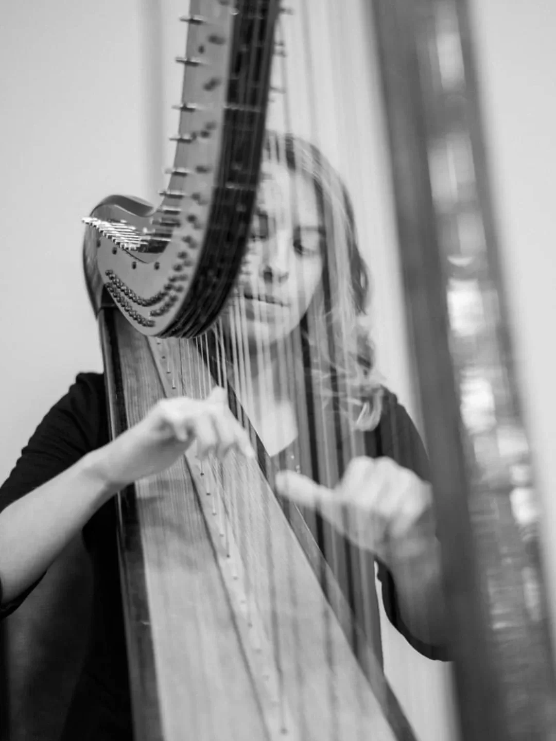 A woman is playing a harp, viewed through a glass surface, creating a reflective and artistic visual effect in black and white.