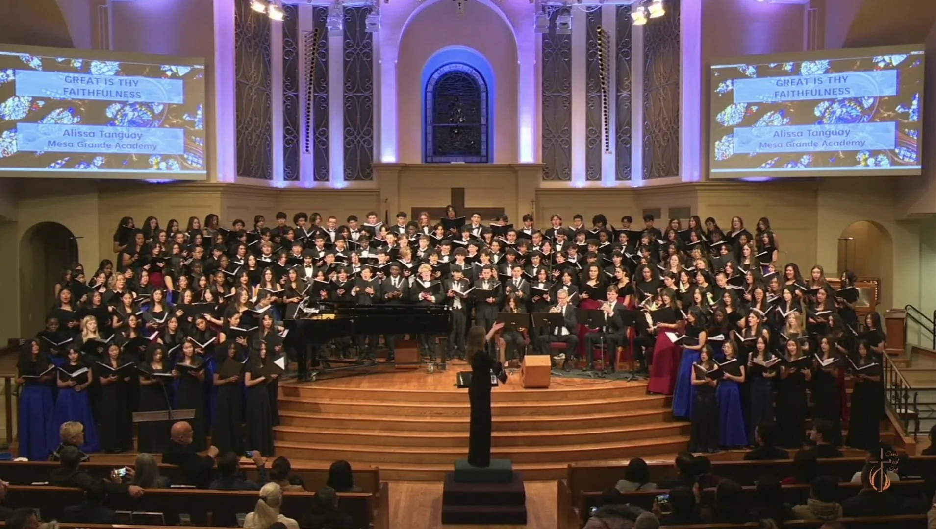 A large choir performing on stage in a church or concert hall, with a conductor leading them. There are two large screens displaying the title 'Great is Thy Faithfulness' and the performer’s name, Alissa Tanguay from Mesa Grande Academy. Audience members are seated in front, watching the performance.