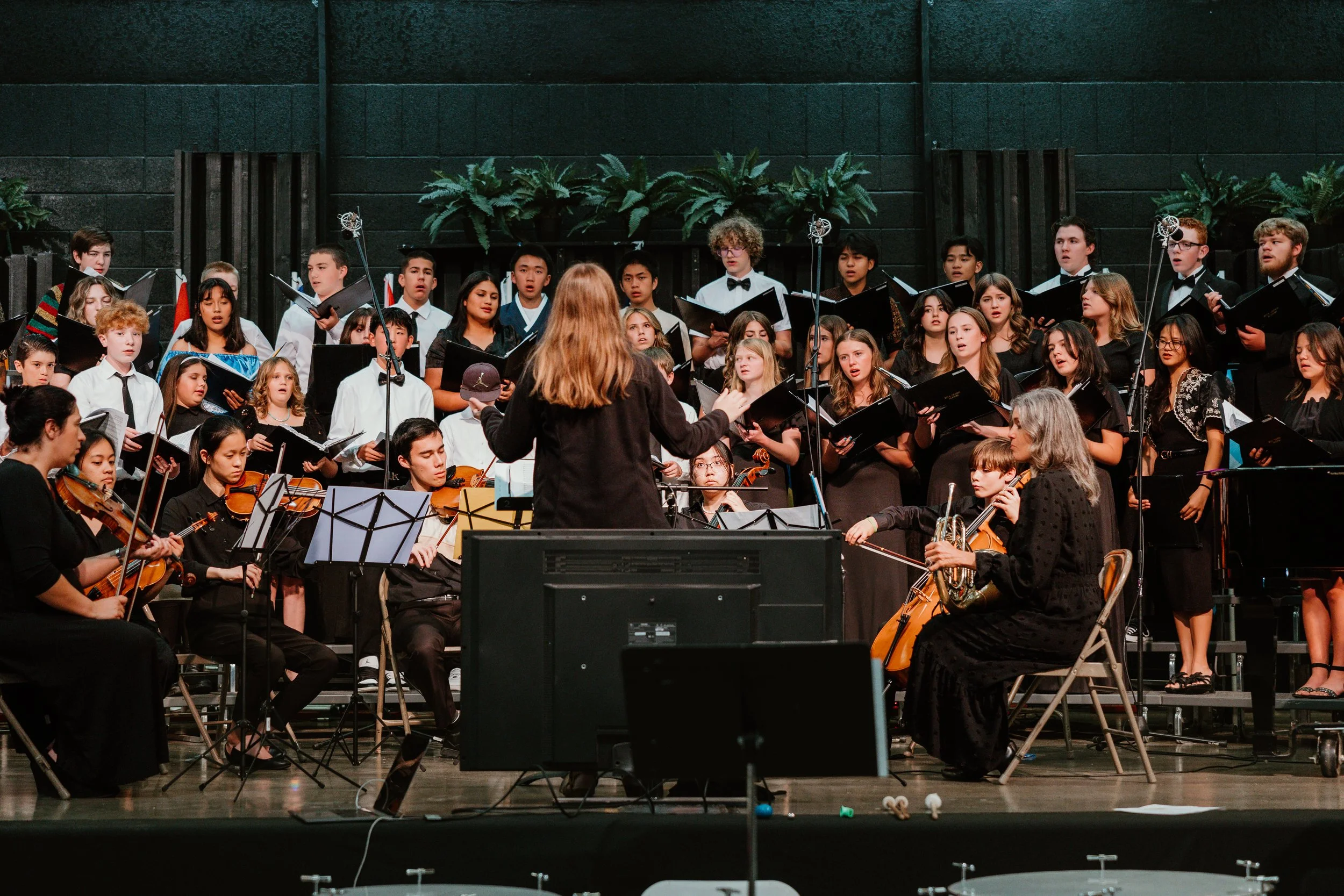 A symphony orchestra and choir performing on stage, with the conductor leading them. The orchestra includes violinists and cellists, and the choir consists of singers dressed in black, standing behind the orchestra.