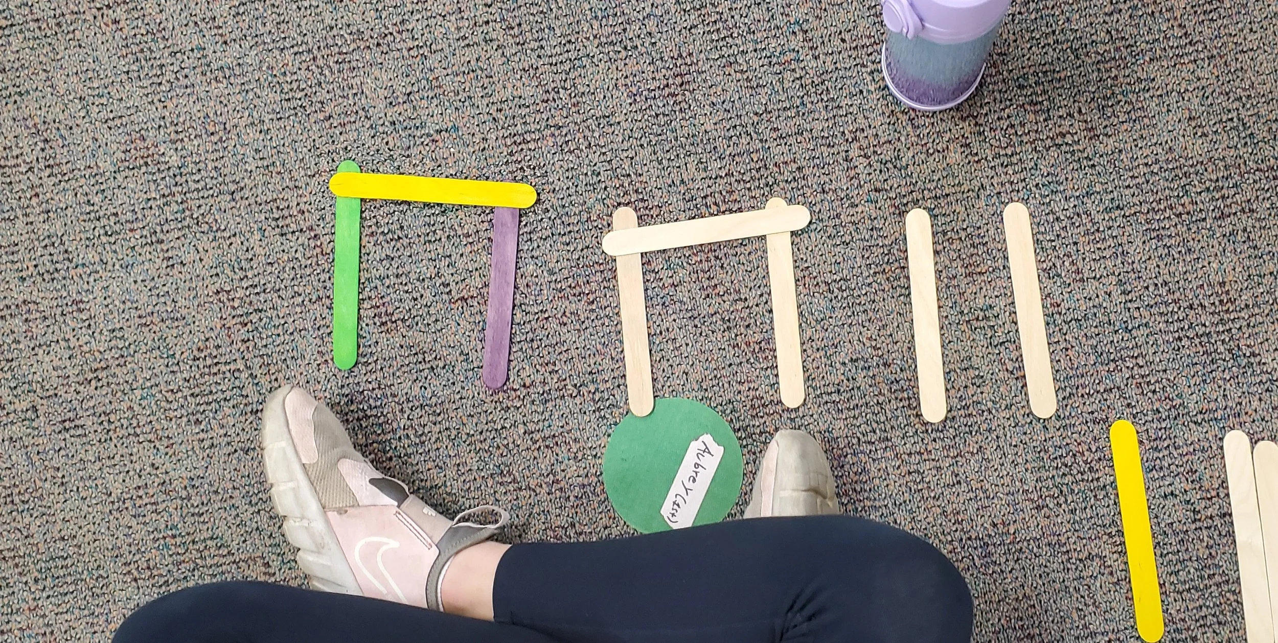 Colorful popsicle sticks arranged on a carpeted floor with a person's foot and leg visible in the foreground, and a purple and green tumbler in the background.