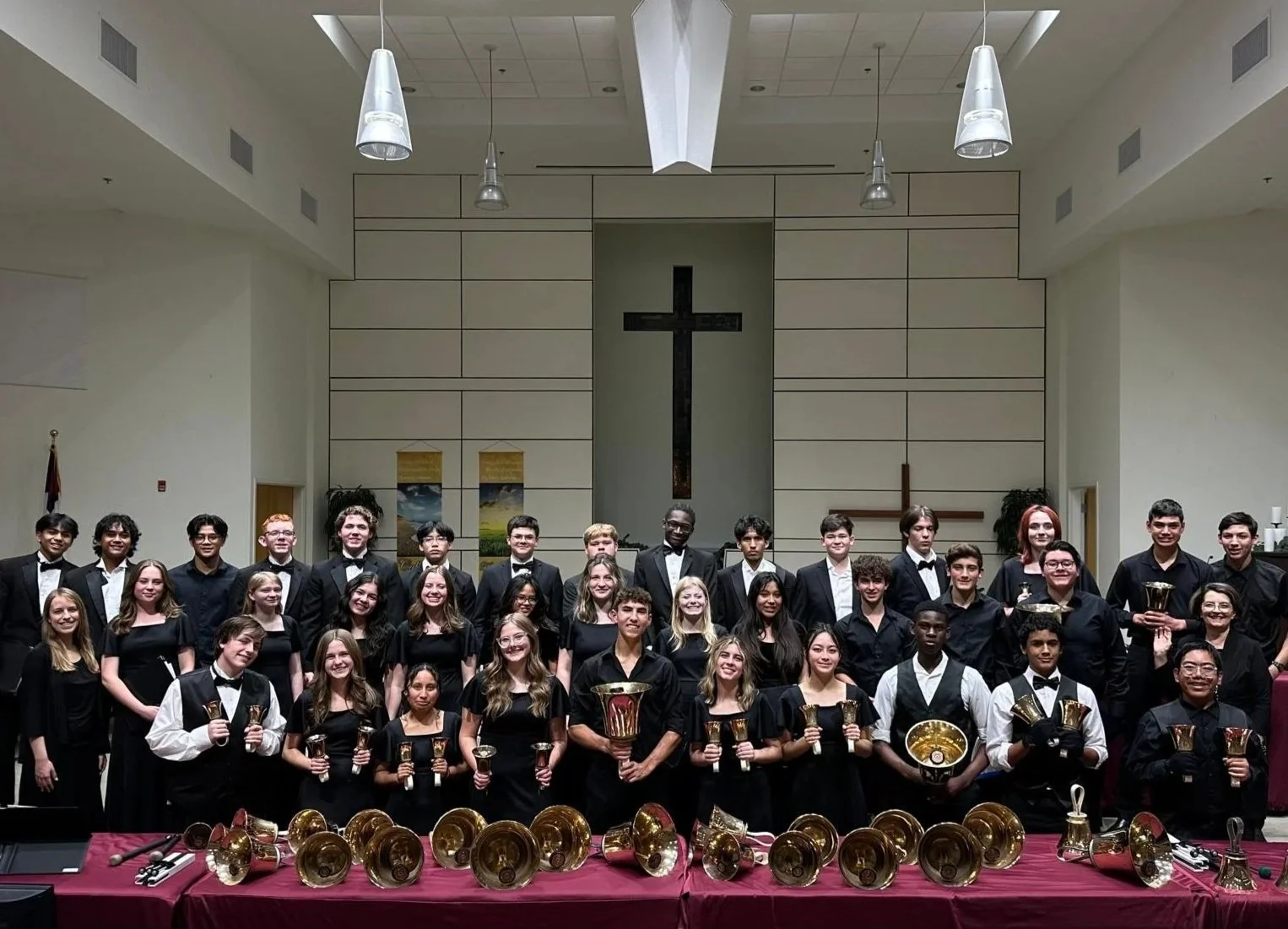 Group of young musicians dressed in black formal attire, holding musical instruments, and standing on a stage inside a church with a large cross on the wall behind them. The stage has a red tablecloth with many small bells placed on it.