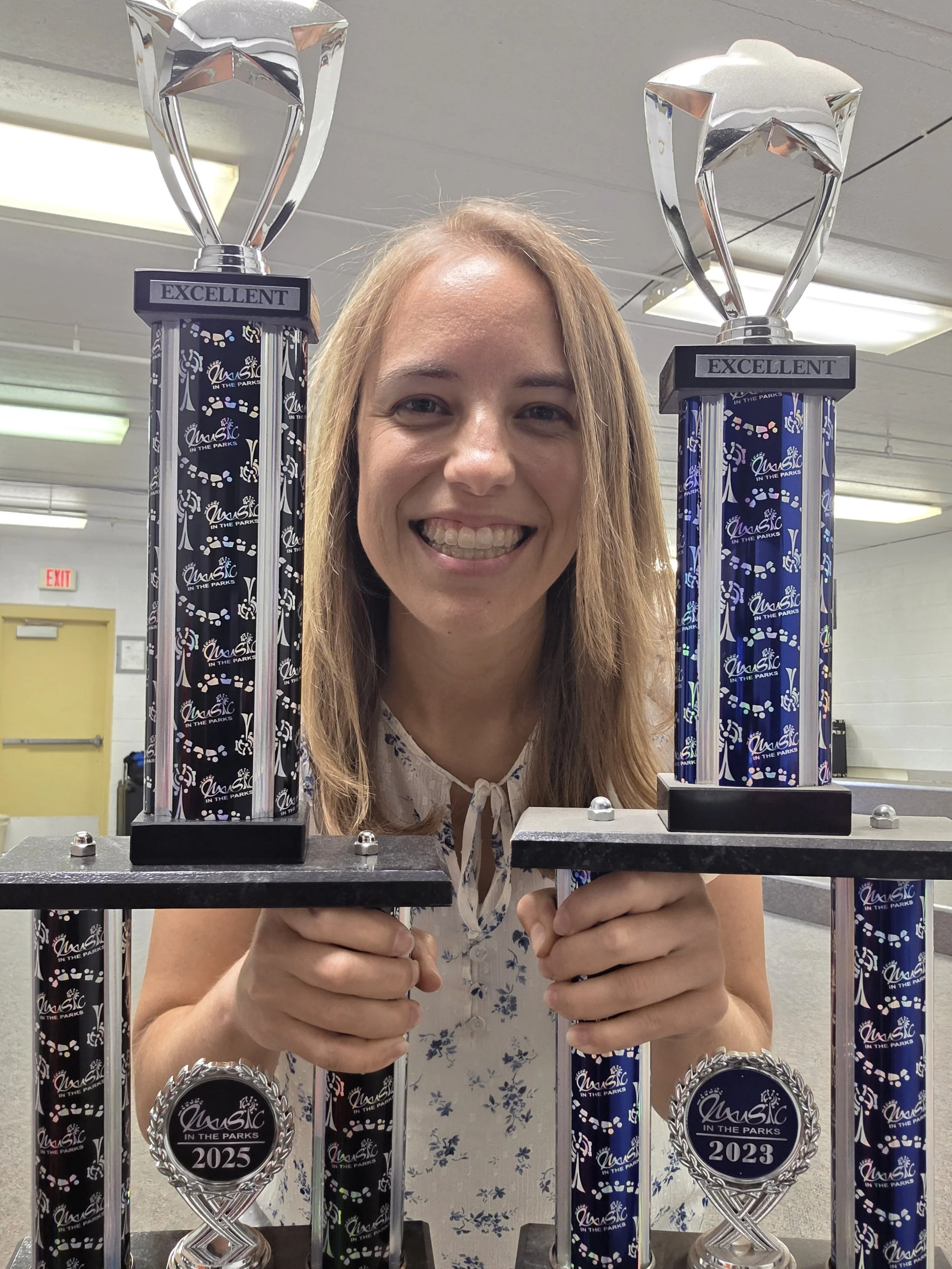 A woman smiling and holding two large trophies at a park event, with medals labeled 2023 and 2025 in front of her.
