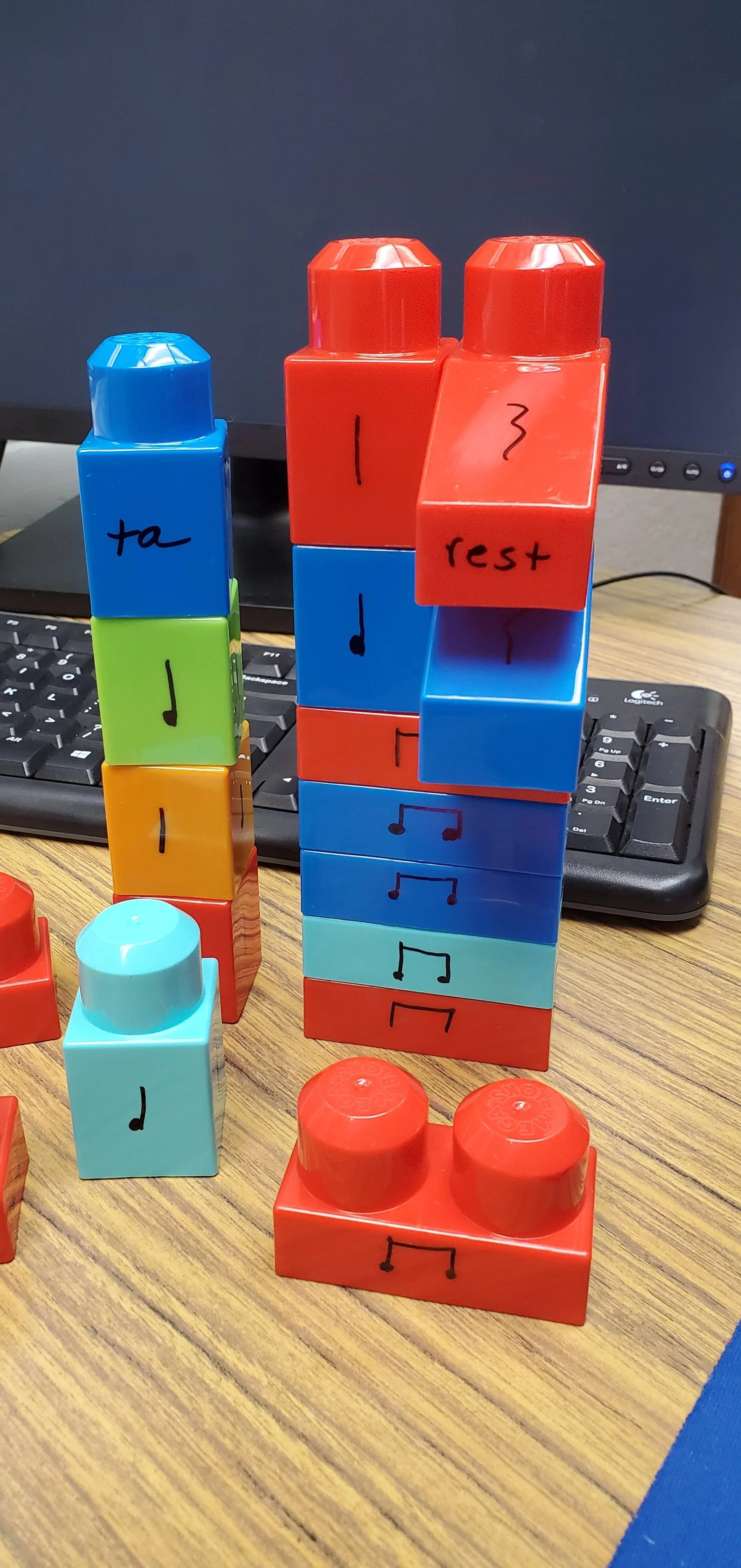 Colorful stacking blocks on a wooden desk in front of a computer keyboard and monitor, with musical notes and text written on them.