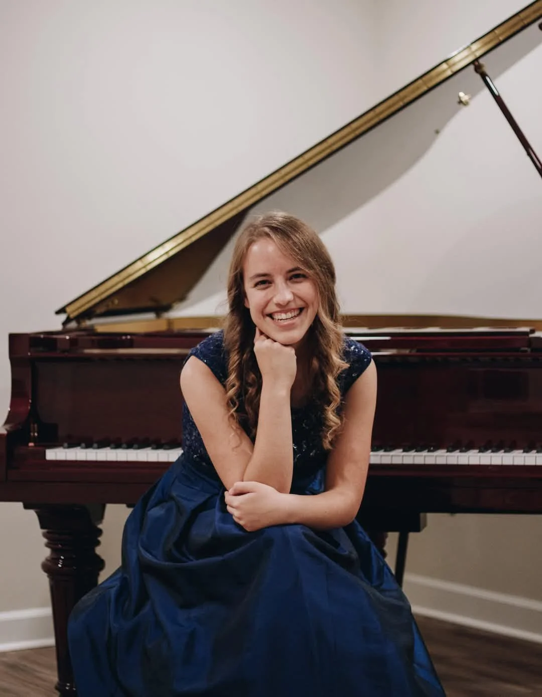 A young woman with long curly brown hair, wearing a sleeveless dark blue dress, sitting in front of a grand piano, smiling and resting her chin on her right hand, in a room with a plain wall.