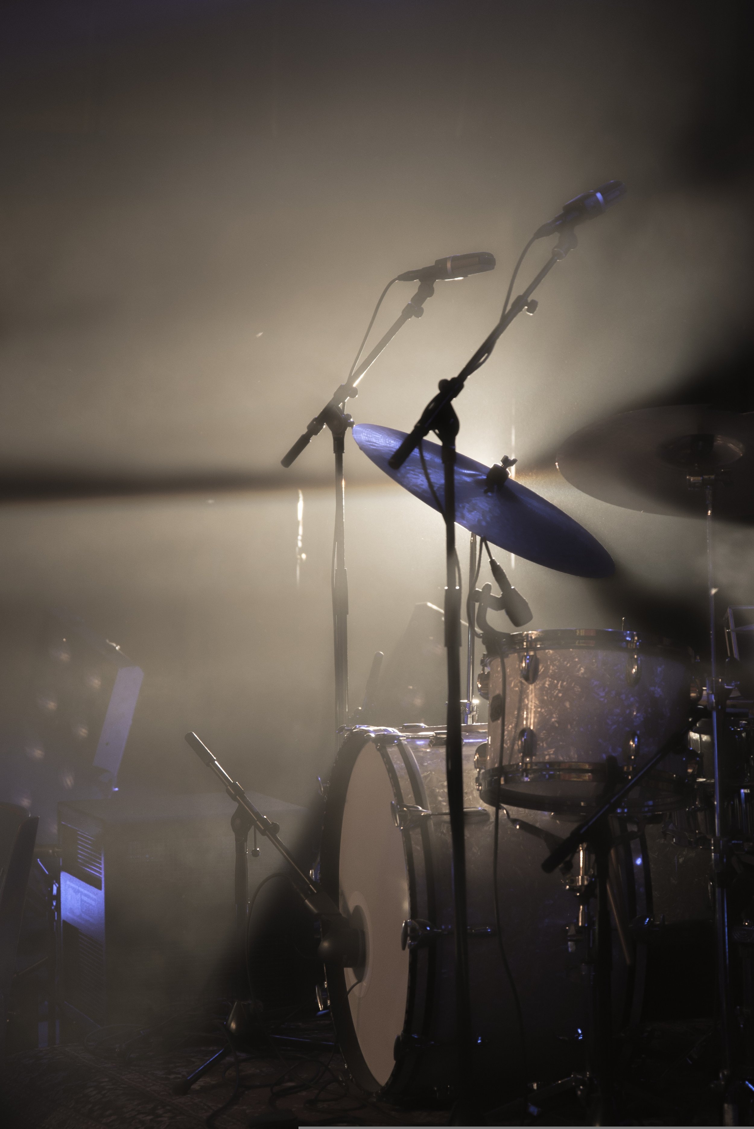 A drum set with cymbals and microphones on stands, illuminated with stage lighting, and enveloped in stage fog or smoke.