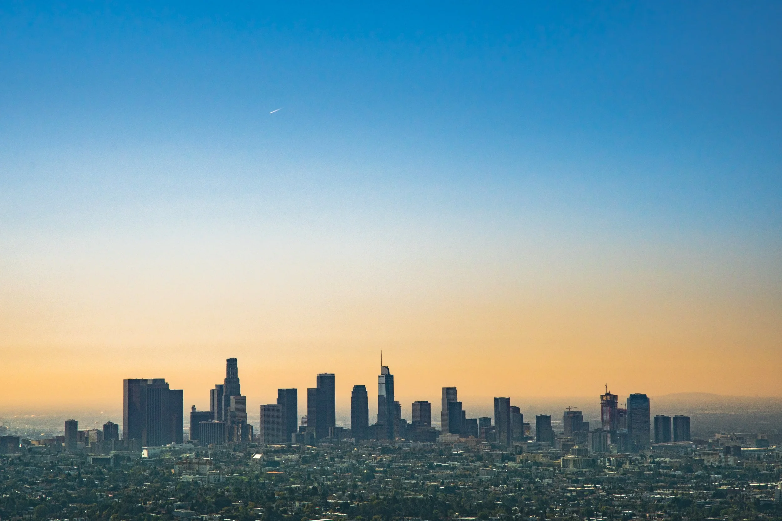Skyline of Los Angeles with tall skyscrapers and a clear blue sky with a faint streak of airplane contrail.