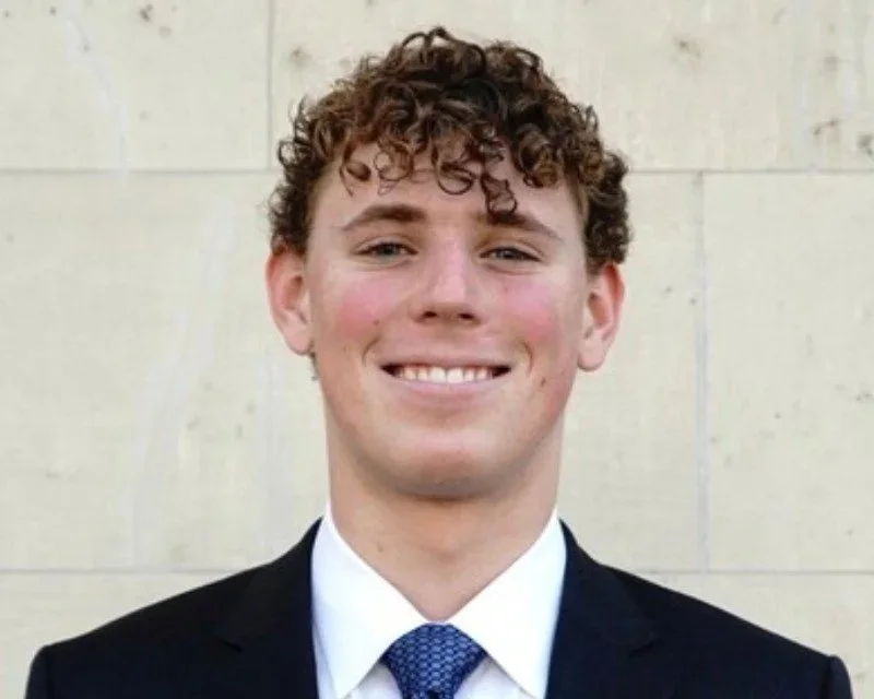 Young man with curly brown hair wearing a black suit, white shirt, and blue tie, smiling in front of a beige wall.