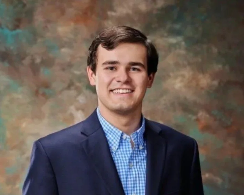 A young man with brown hair wearing a blue blazer and a blue checkered shirt, smiling for a portrait against a painterly brown and green background.