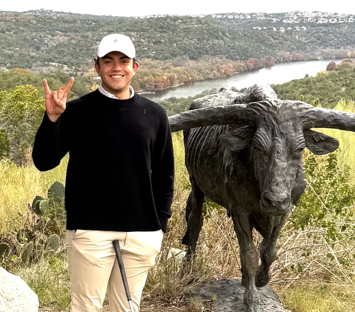 A young man in a black sweater and white pants making a rock on hand sign next to a buffalo sculpture in a natural landscape with a river and hills in the background.