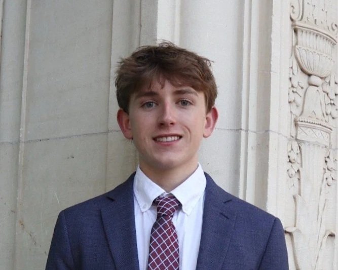 Young man in a dark blue suit, white shirt, and patterned tie standing outdoors next to a stone building with decorative carvings.