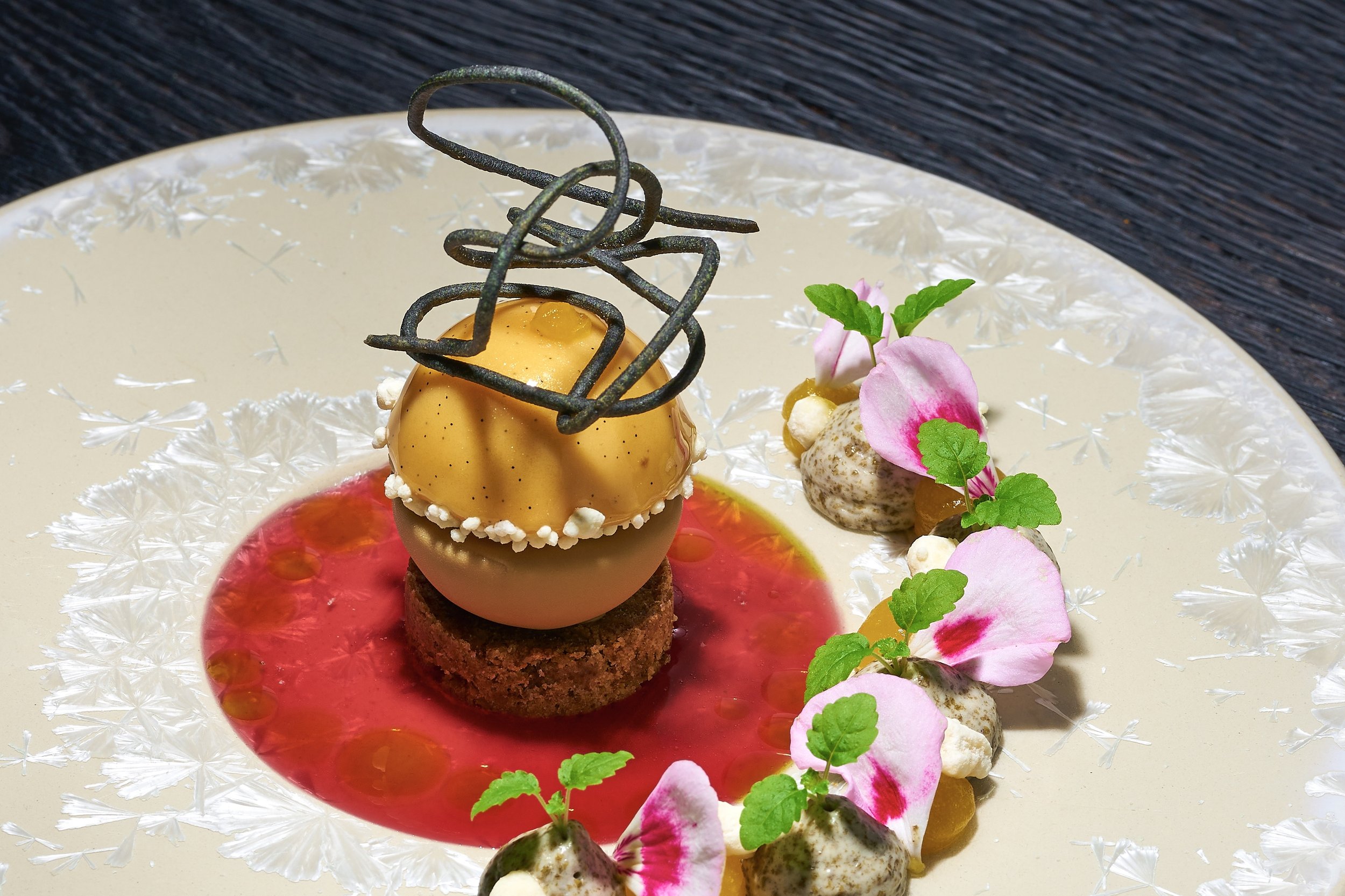 Elegant plated dessert with a yellow caramel-glazed sphere on a chocolate cookie, topped with a black decorative chocolate piece, surrounded by a red sauce with small droplets, pink edible flower petals, green leaves, and dollops of cream or mousse.