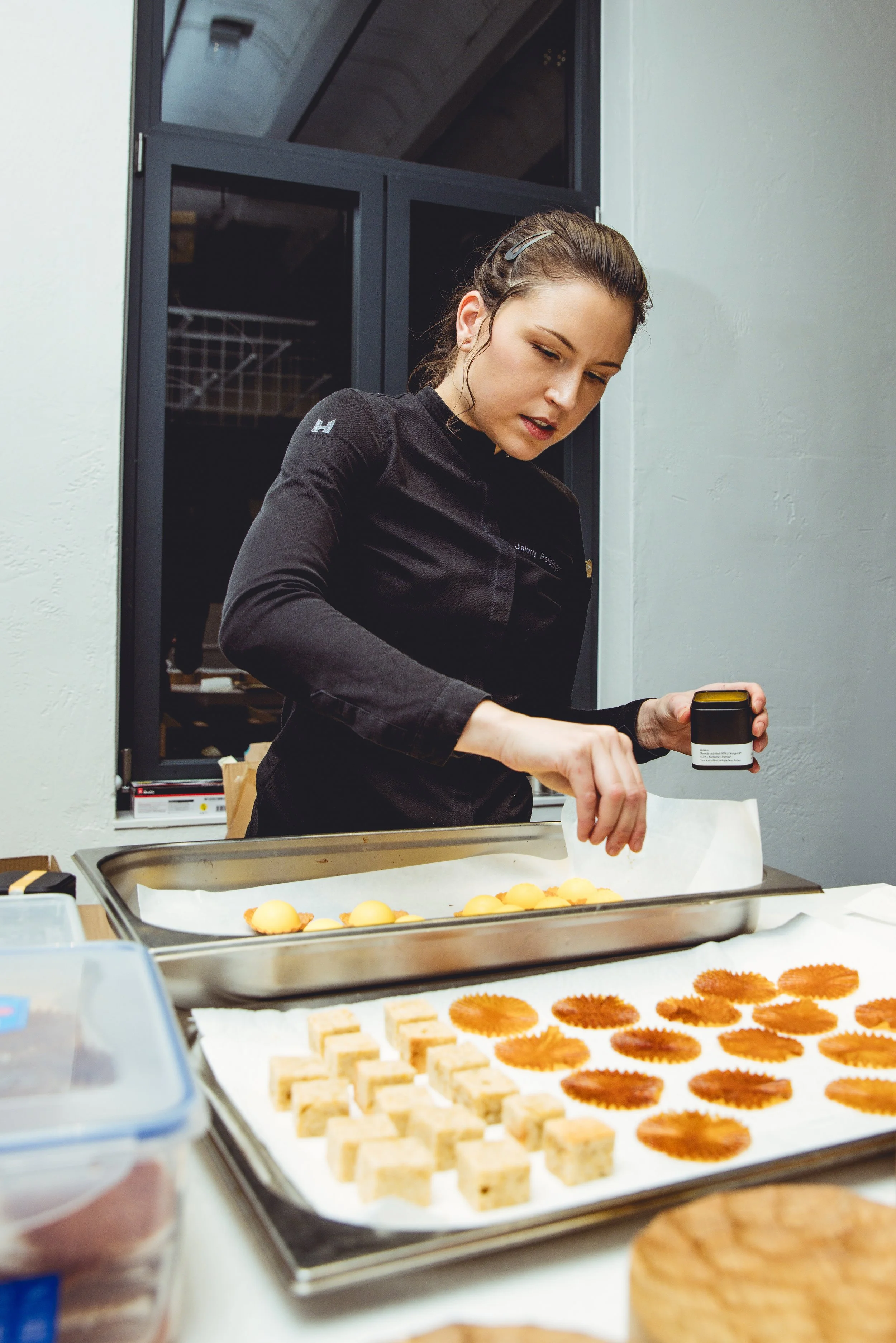 Jaimy Reisinger in a black chef's coat sprinkling seasoning on pastries at a baking station.