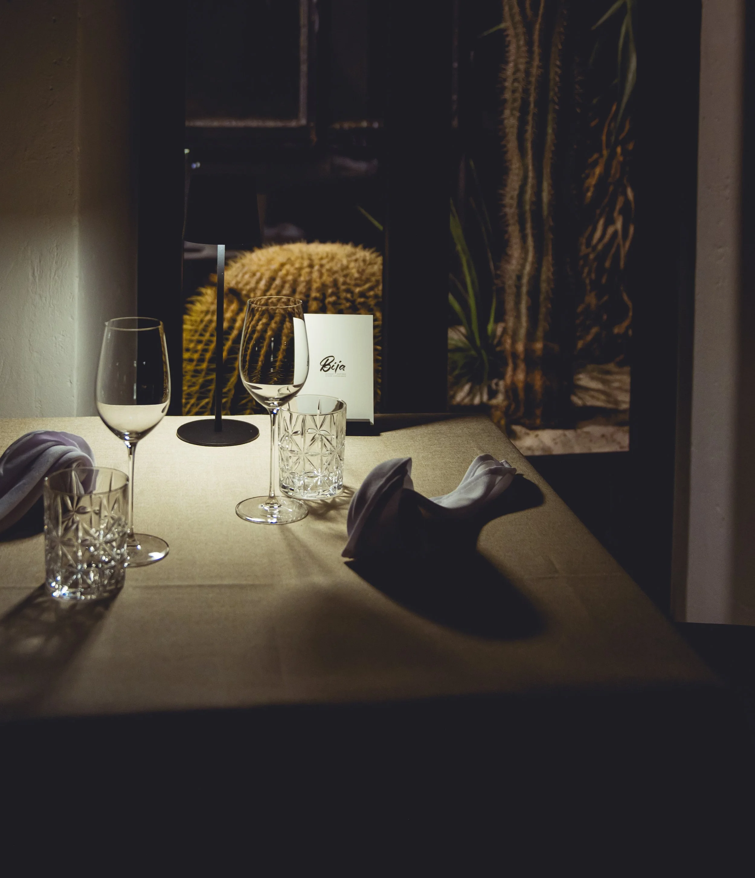 A dimly lit restaurant table set with wine glasses, water glasses, and napkins, with a small black lamp, a menu, and a mirror reflecting a cactus plant and a fluffy decor piece.