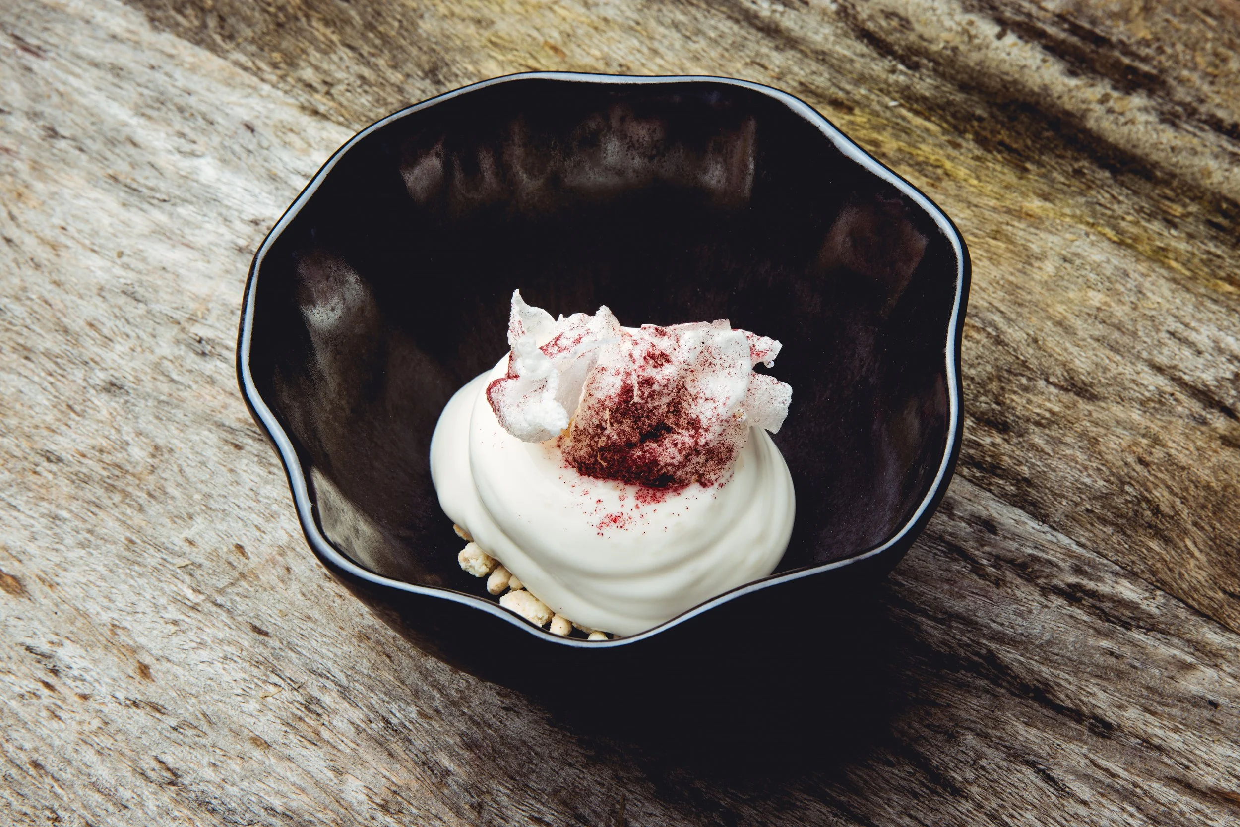 A black bowl contains a dessert with a dollop of white cream, topped with white cotton candy and a dusting of red powder, on a wooden surface