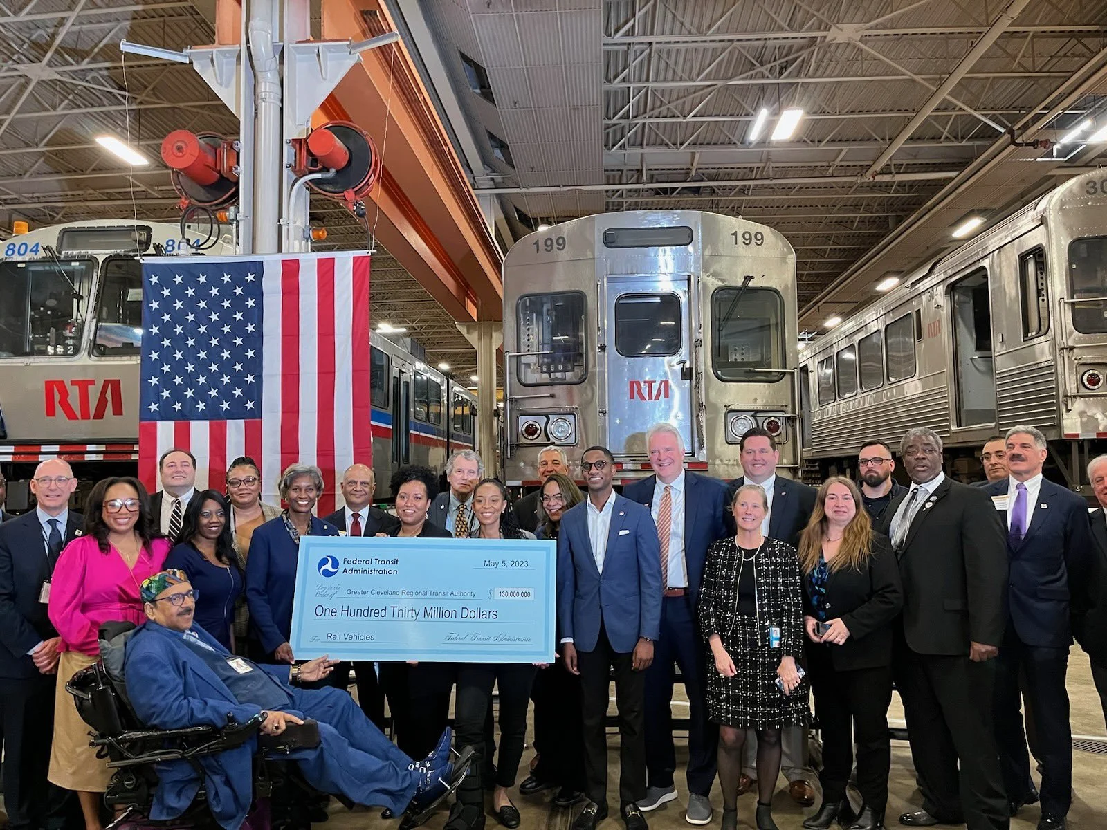 Group of people holding a large check inside a train depot with trains and an American flag in the background.