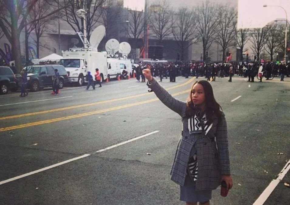 A young woman standing on a city street with her arm raised, possibly holding a phone. In the background, there are vehicles, including press vans with satellite dishes, and a crowd of people on the sidewalk, suggesting a public event or protest.