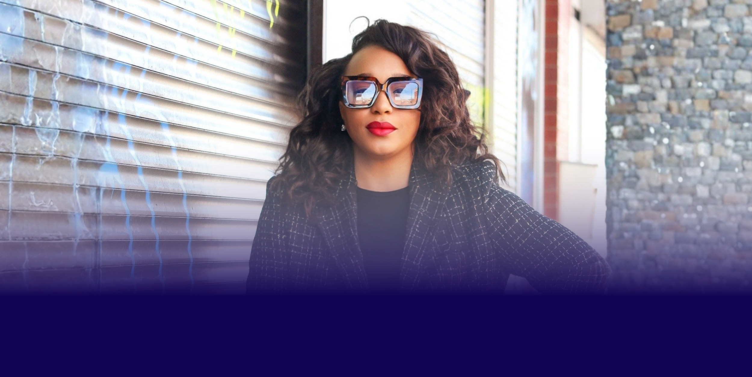 A woman with curly dark hair wearing large sunglasses and a black blazer with a checkered pattern is sitting outdoors against a metal and stone wall.