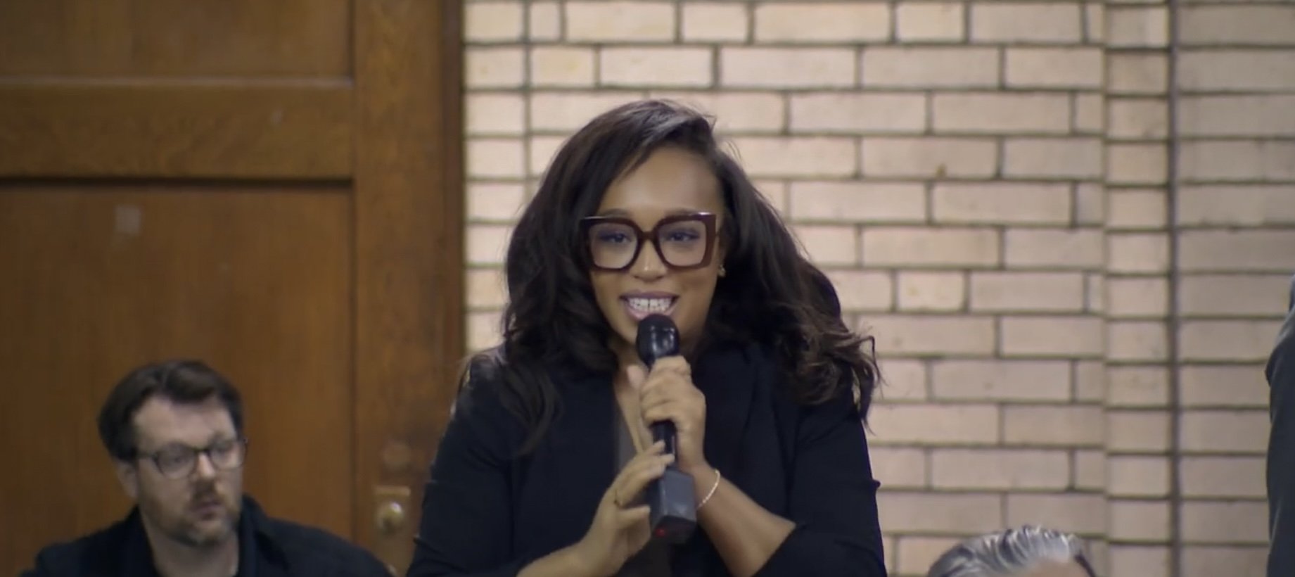 Woman with curly hair and glasses smiling and speaking into a microphone during a meeting or event, with a brick wall background.