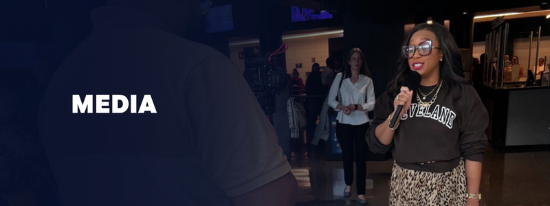 A woman in glasses holding a microphone speaking at an indoor event with a media overlay on the left.