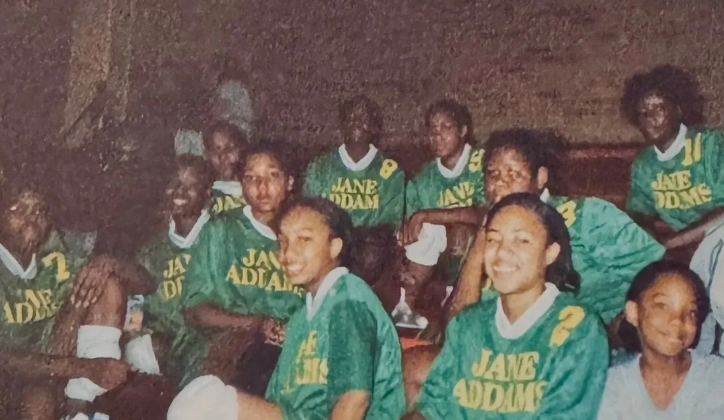 A group of young women, some wearing green sports jerseys with yellow text that reads 'JANE ADDAM' and numbers, sitting together indoors against a textured wall, smiling and posing for the photo.