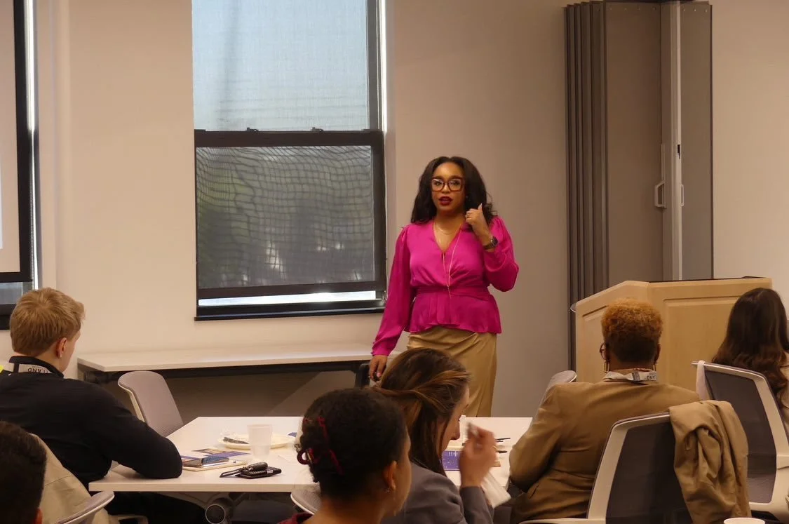 A woman in a pink blouse and beige skirt is standing at the front of a classroom, speaking to a group of students seated at tables. The classroom has large windows with blinds and a classroom podium to her right.