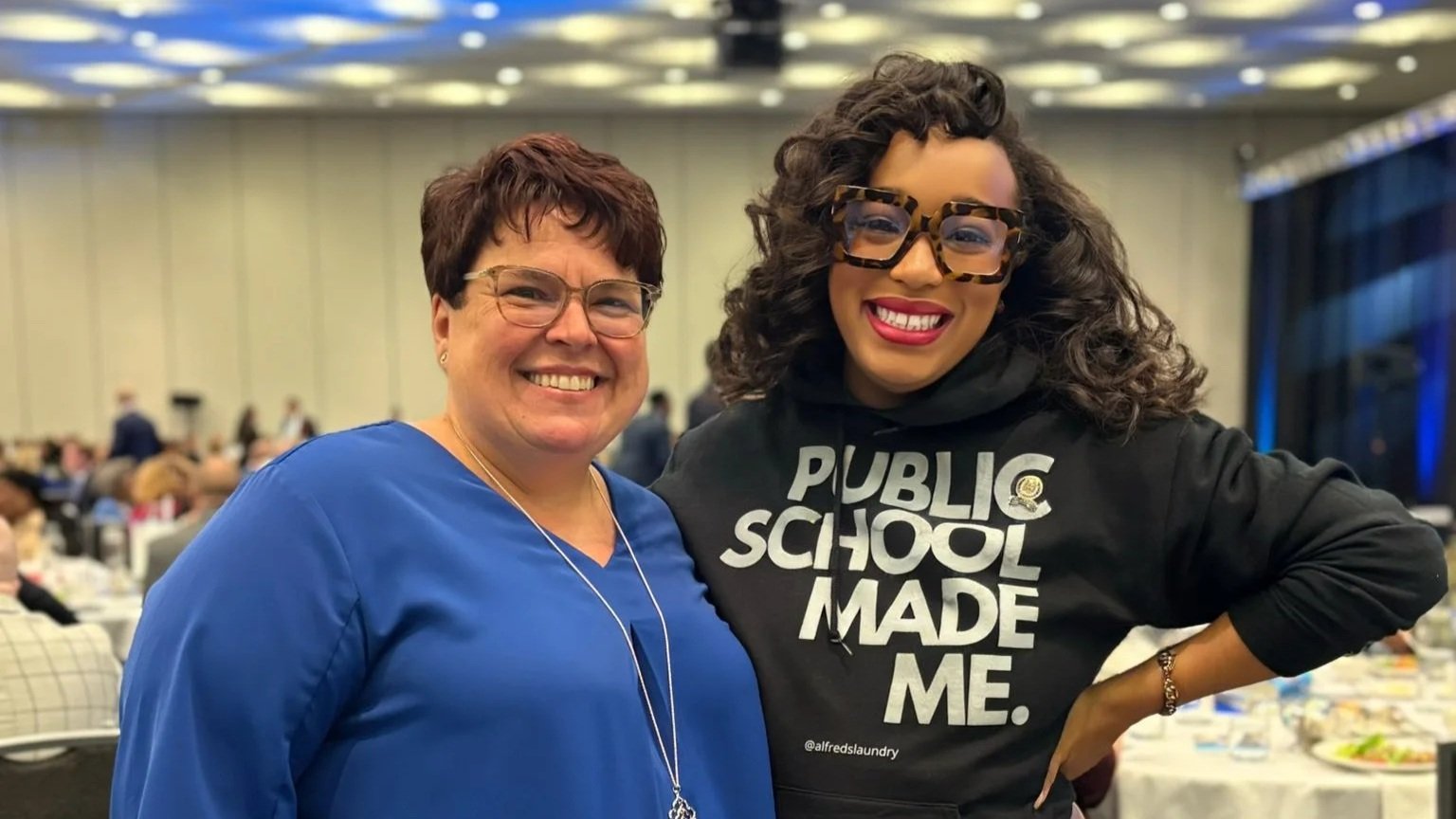 Two women smiling, standing close together in a large conference or banquet hall. One woman is wearing a blue top and glasses, the other is wearing a black hoodie that says 'Public School Made Me' and large tortoiseshell glasses.