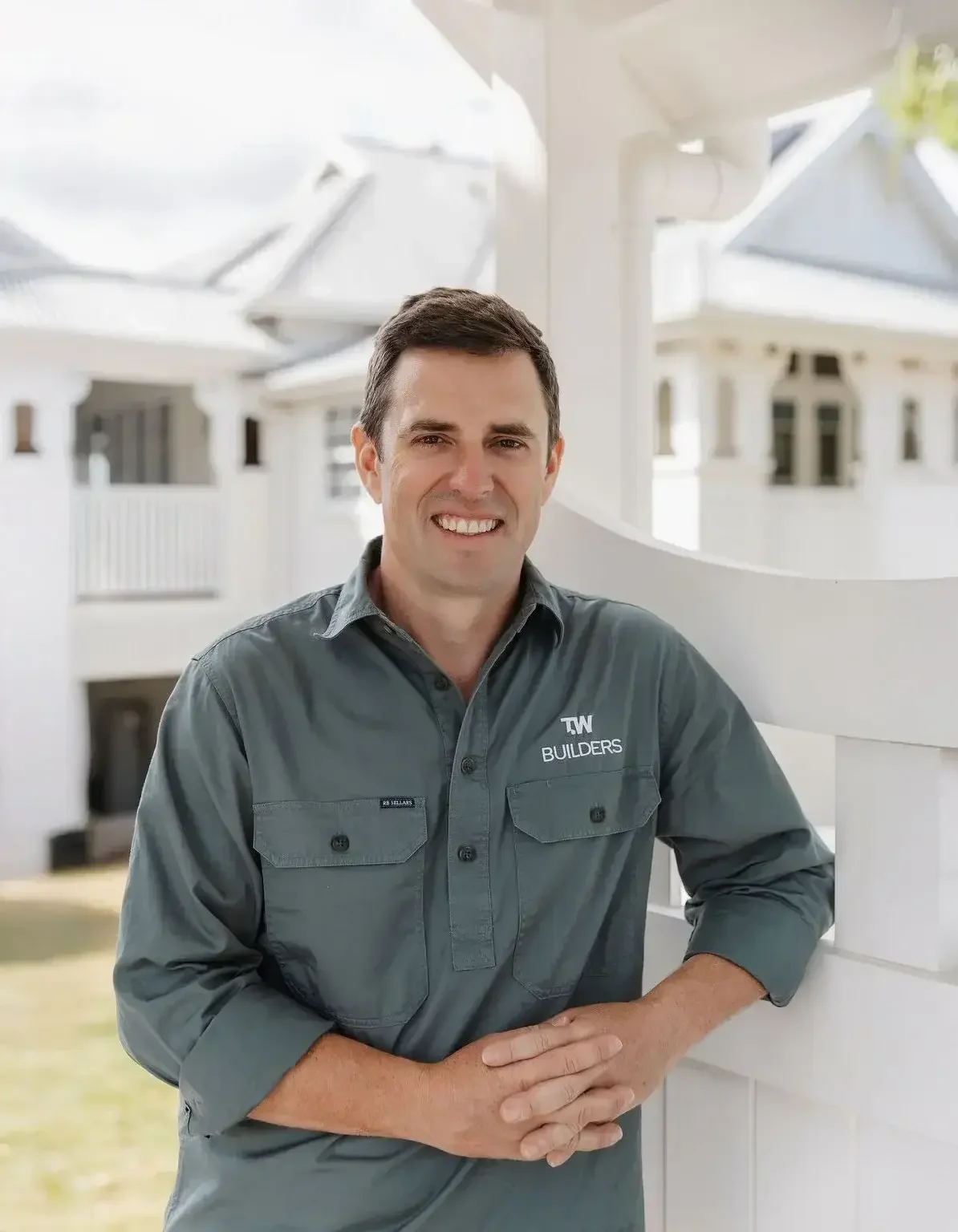 A man in a gray work shirt with 'TW BUILDERS' logo, smiling and standing outdoors near a white porch, with houses and sunny sky in the background.