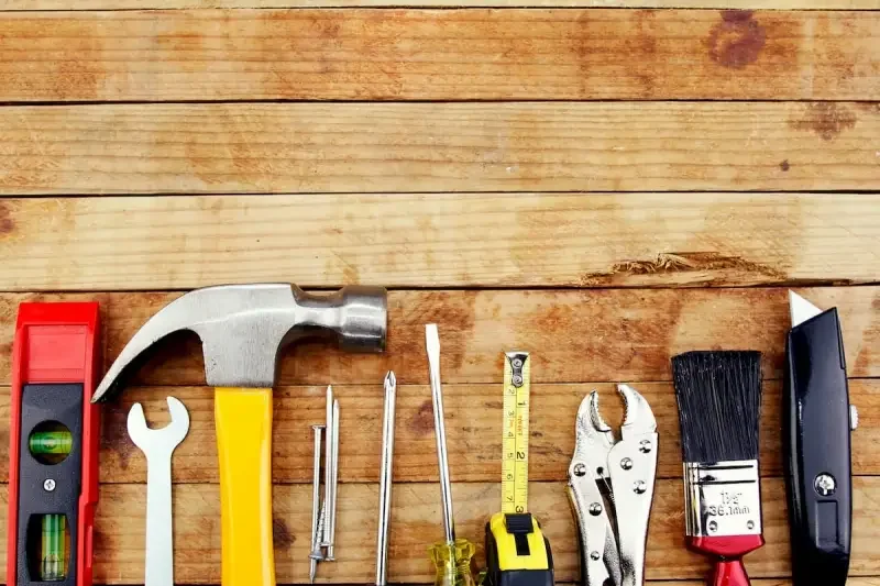 Collection of various hand tools on a wooden surface, including a level, wrench, hammer, screwdrivers, measuring tape, pliers, paintbrush, and utility knife.