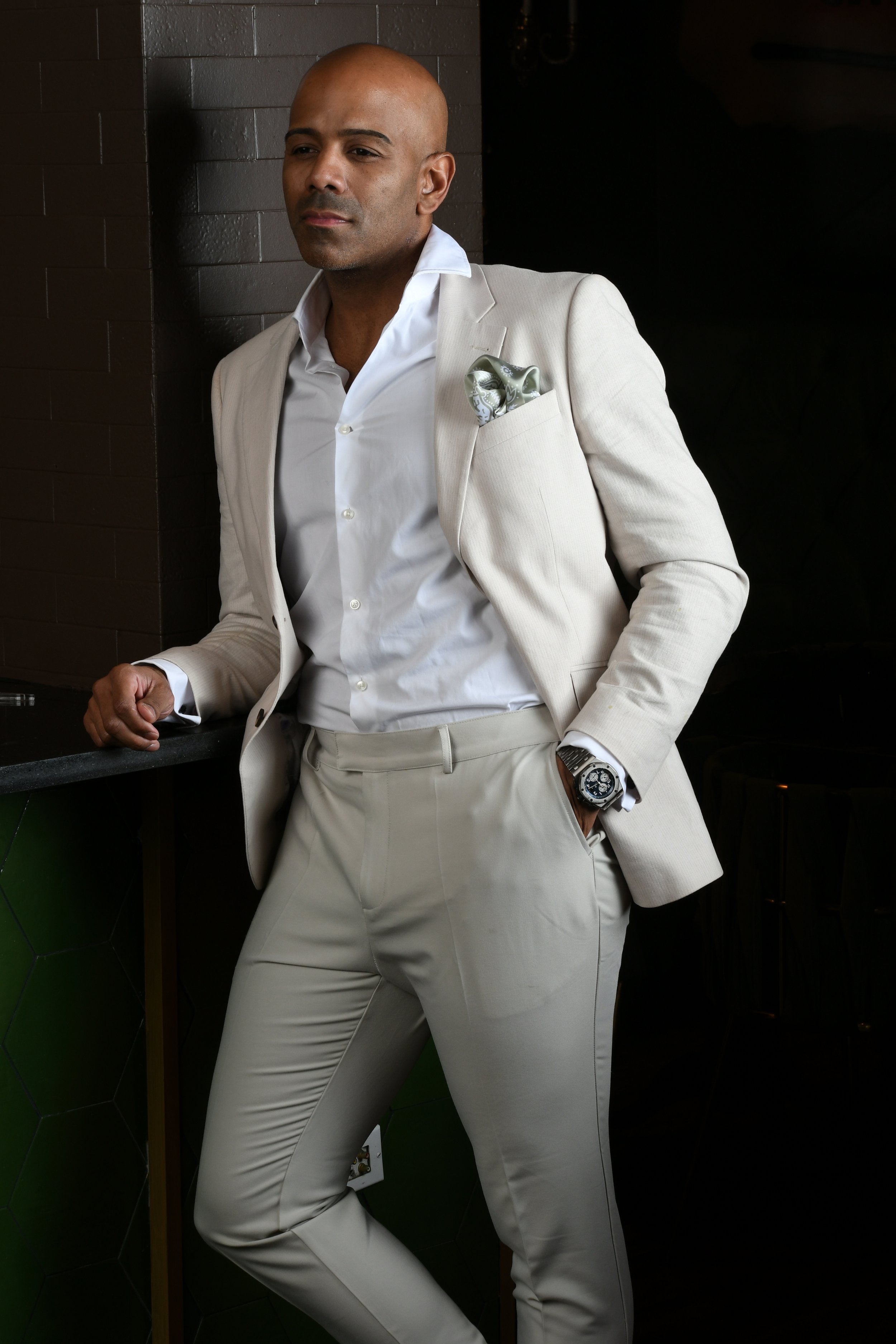 Sanjay Laforest in a cream suit no belt with a patterned pocket square, standing against a dark background.