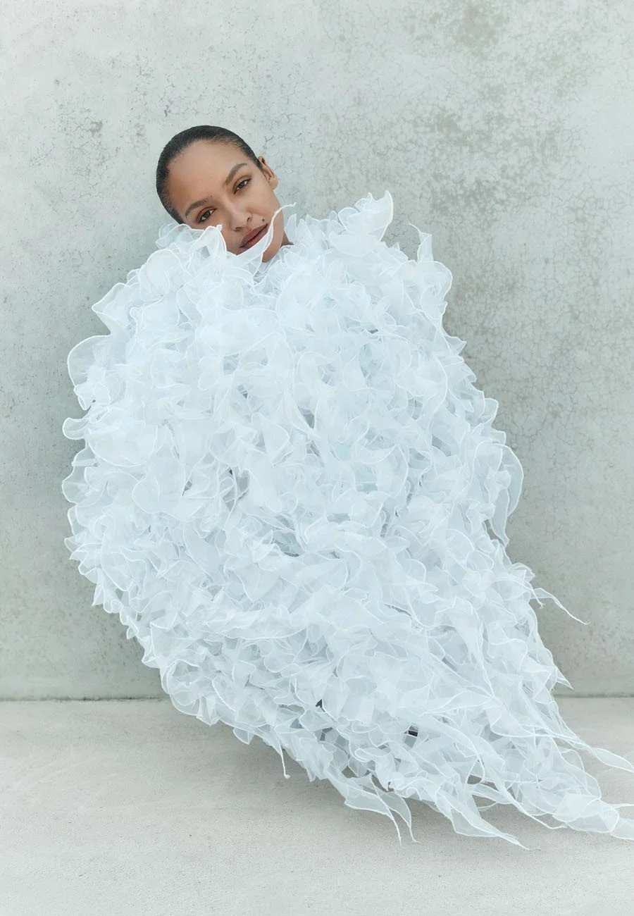 A model with a slick bun hairstyle wearing a white, ruffled, feather-like dress, sitting against a light gray textured wall.