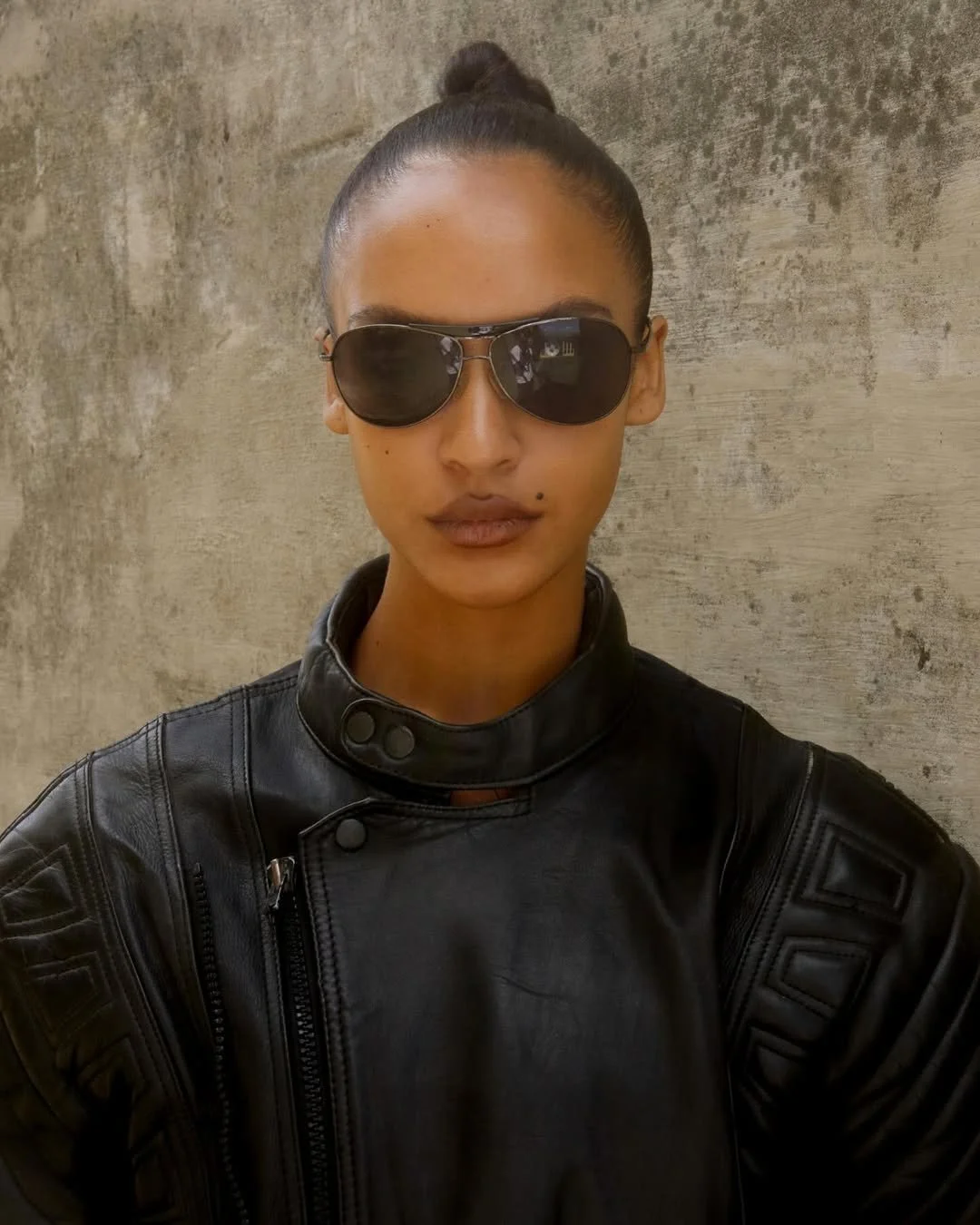 A woman in a black leather jacket and sunglasses standing against a textured wall.