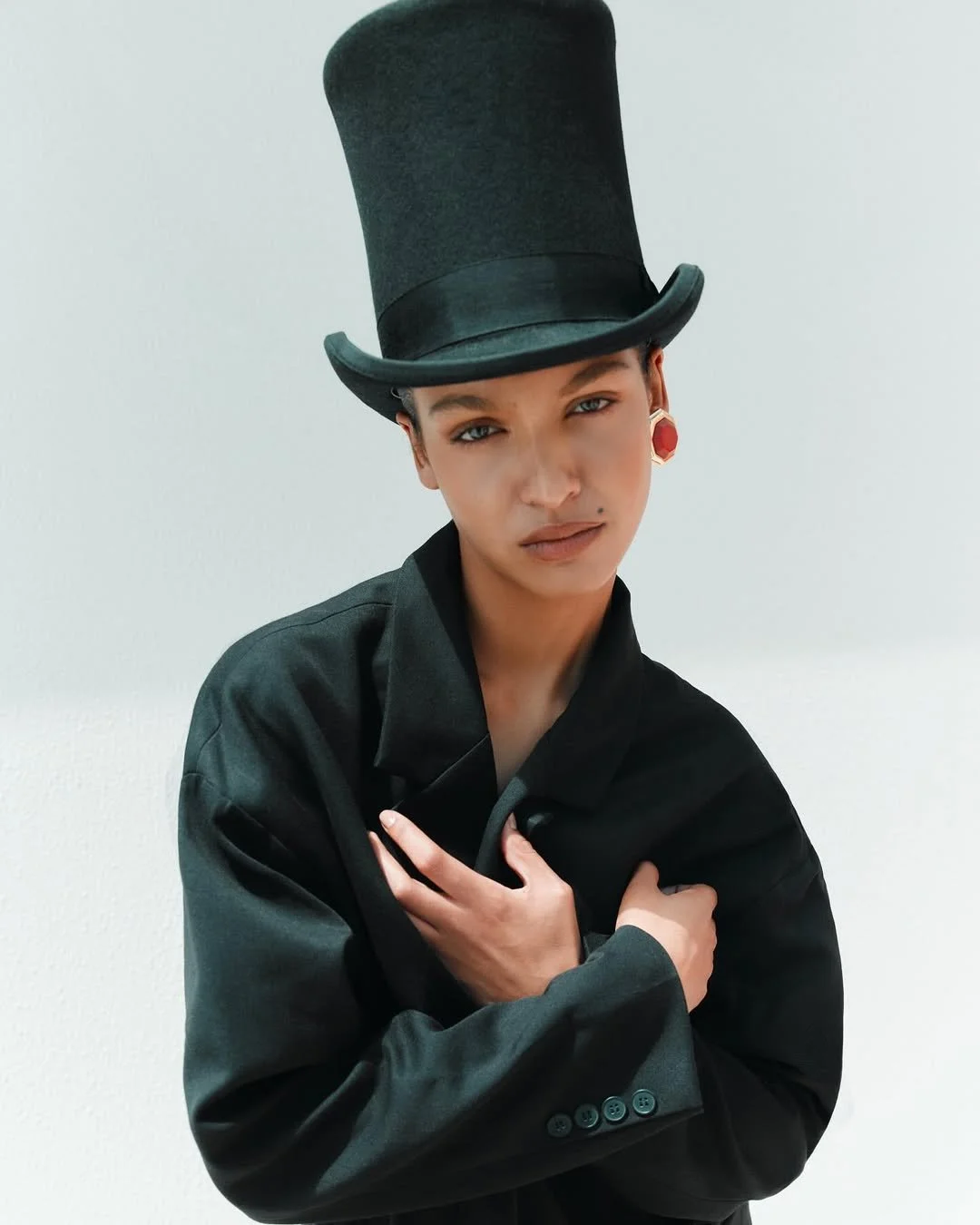 A model wearing a large black top hat, black suit, and red earrings, posing with her arms crossed over her chest against a plain white background.