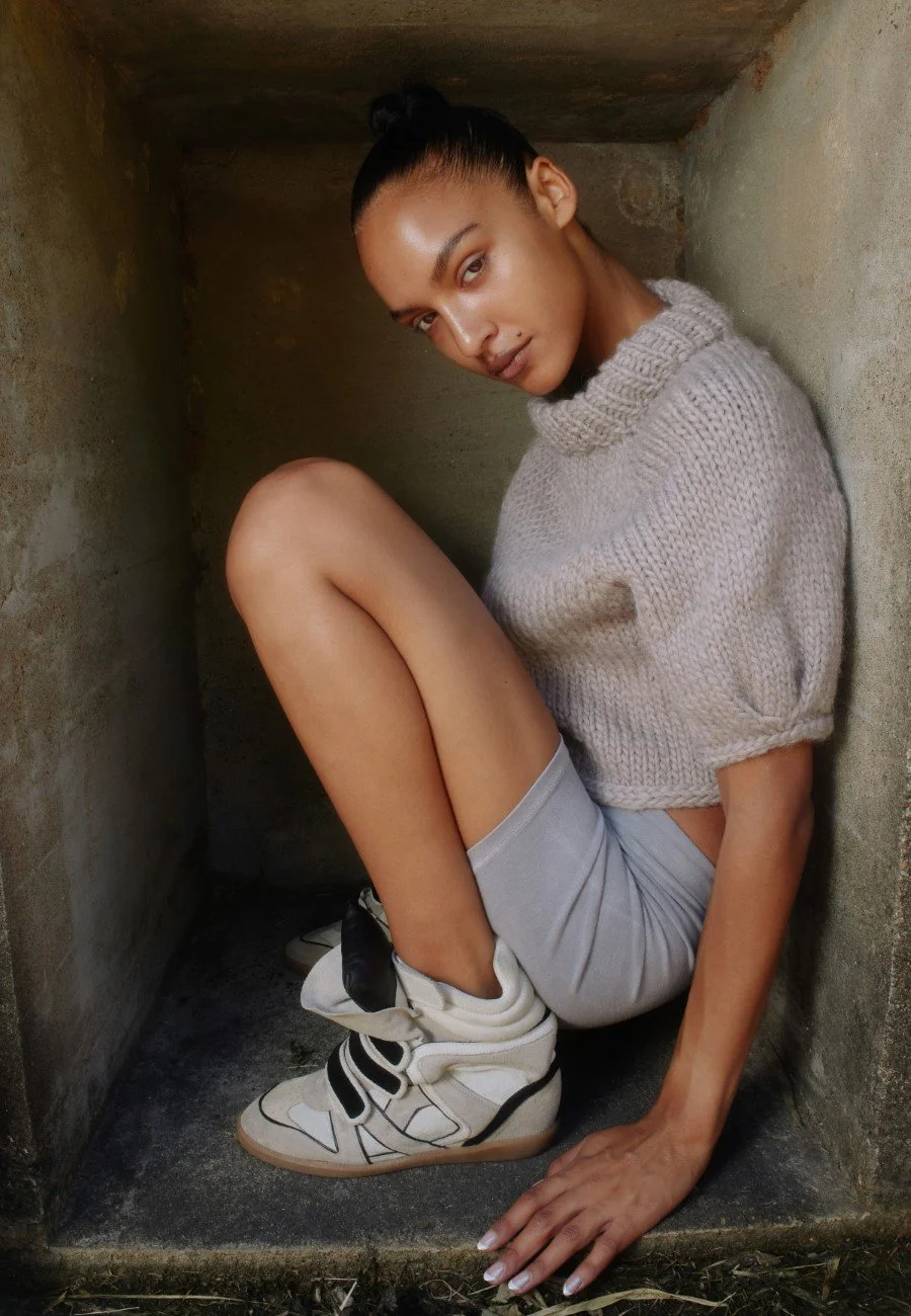 A woman wearing a beige cable knit sweater, beige shorts, and white sneakers, sitting in a small, confined stone space with her knees drawn up and her hair in a bun.