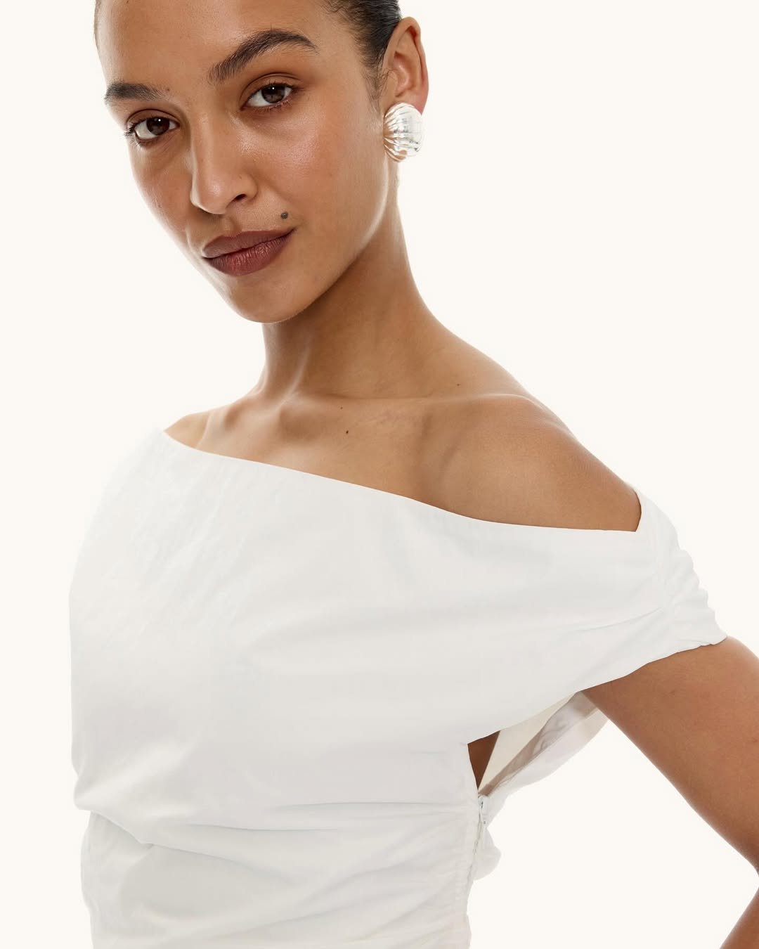 A woman with dark hair pulled back, wearing a white off-shoulder top and large pearl earrings, posing against a plain white background.