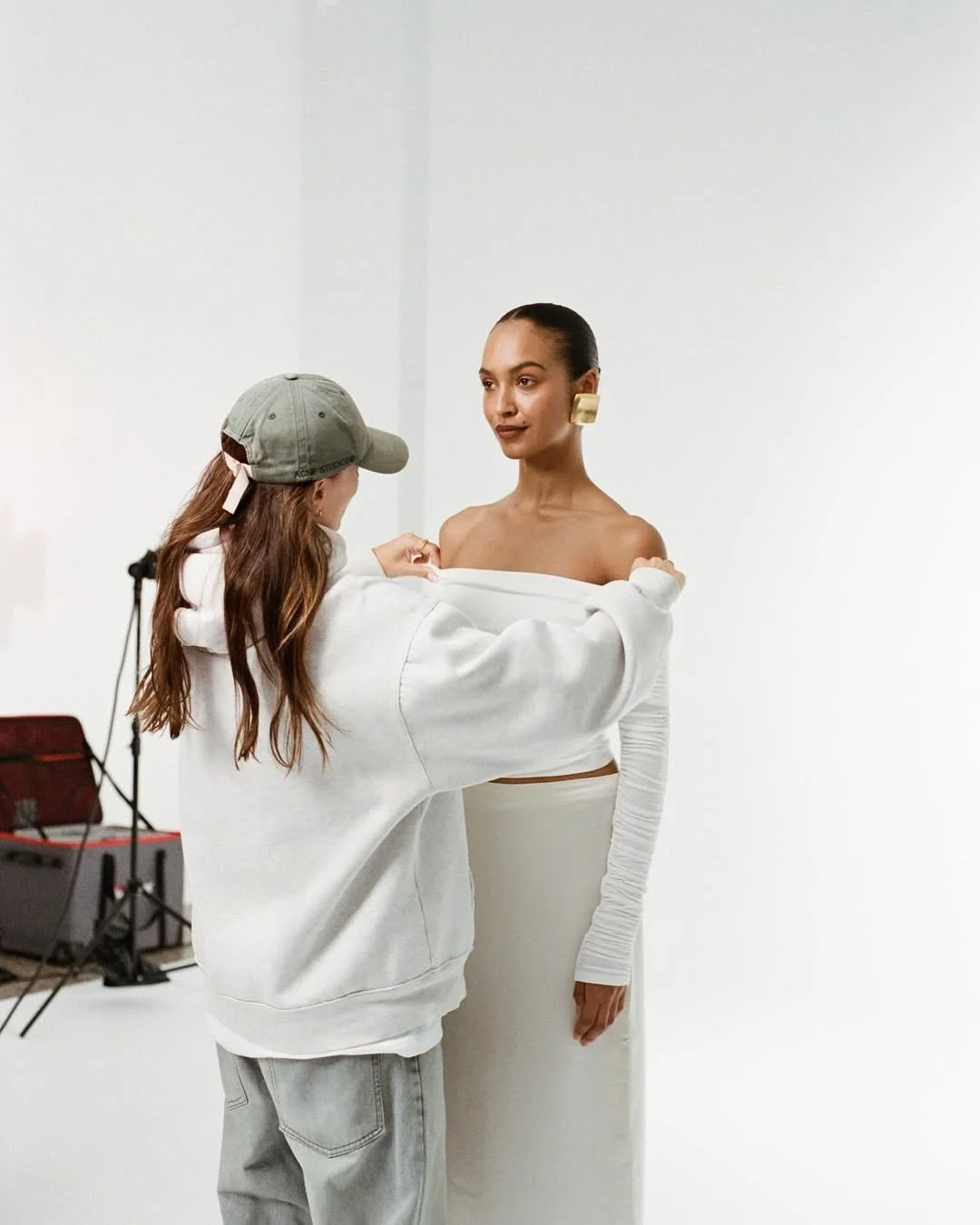A woman in a white off-shoulder top and high-waisted skirt poses for a photoshoot, wearing large gold earrings, while a stylist adjusts her outfit in a bright studio with a plain white background.