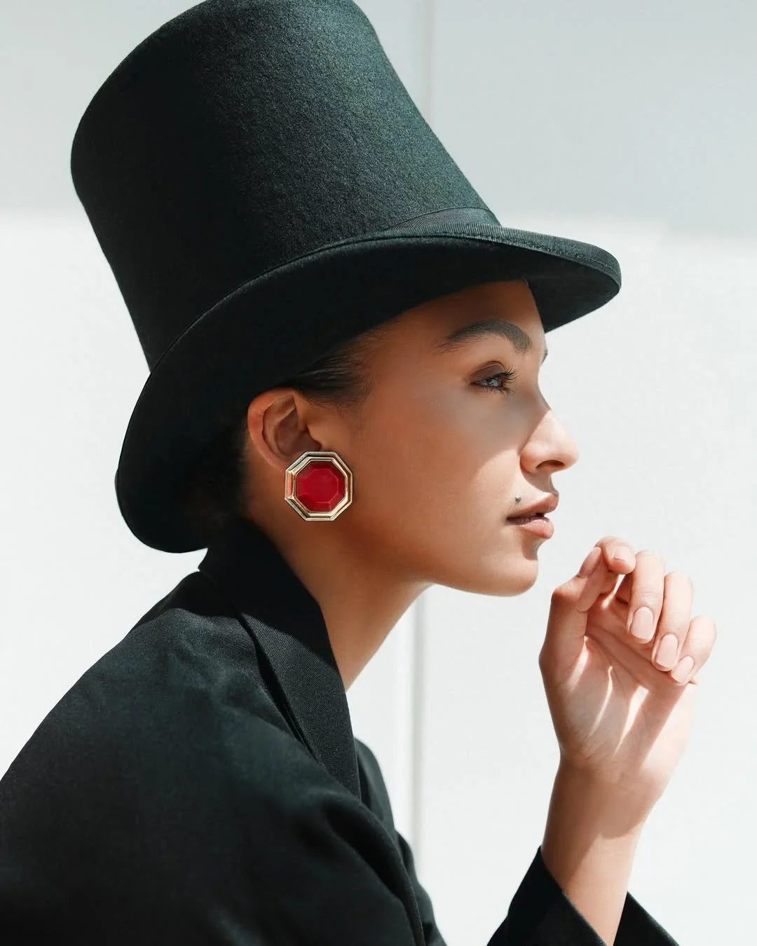 A model with light brown skin wearing a black top hat, large red and silver earrings, and a black blazer, in profile, with her hand near her face against a plain background.