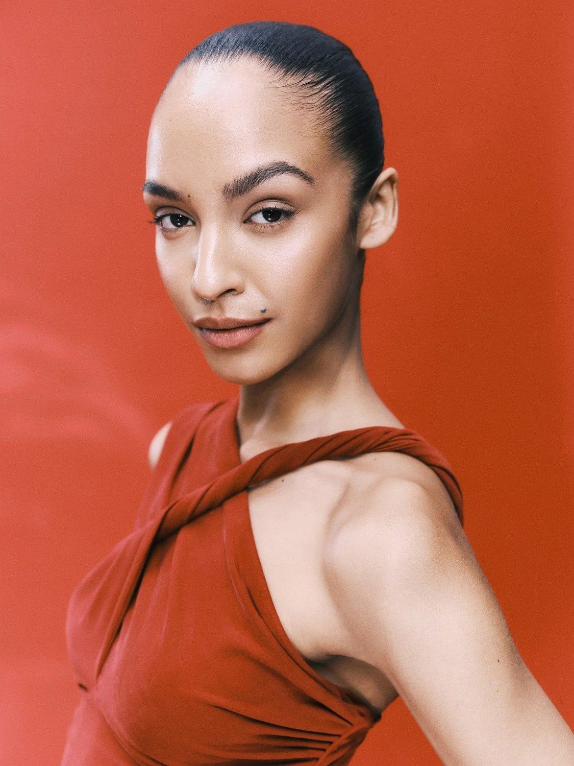 A woman with dark hair styled in a sleek bun, wearing a red dress with cut details on the shoulder, standing against a red background.