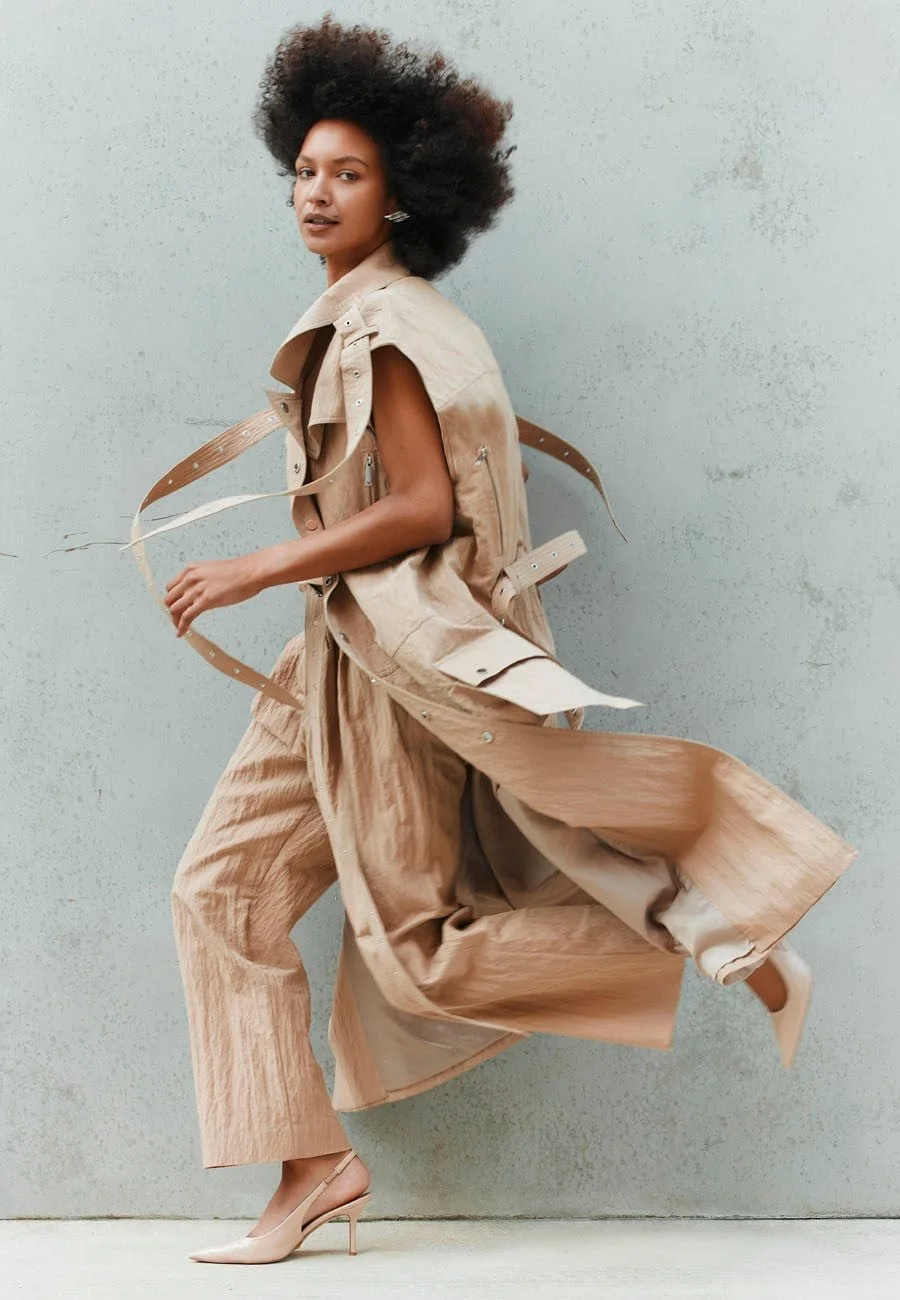 A model with voluminous curly hair wearing a beige trench coat, tan wide-leg pants, and nude high heels mid-stride against a light textured wall.