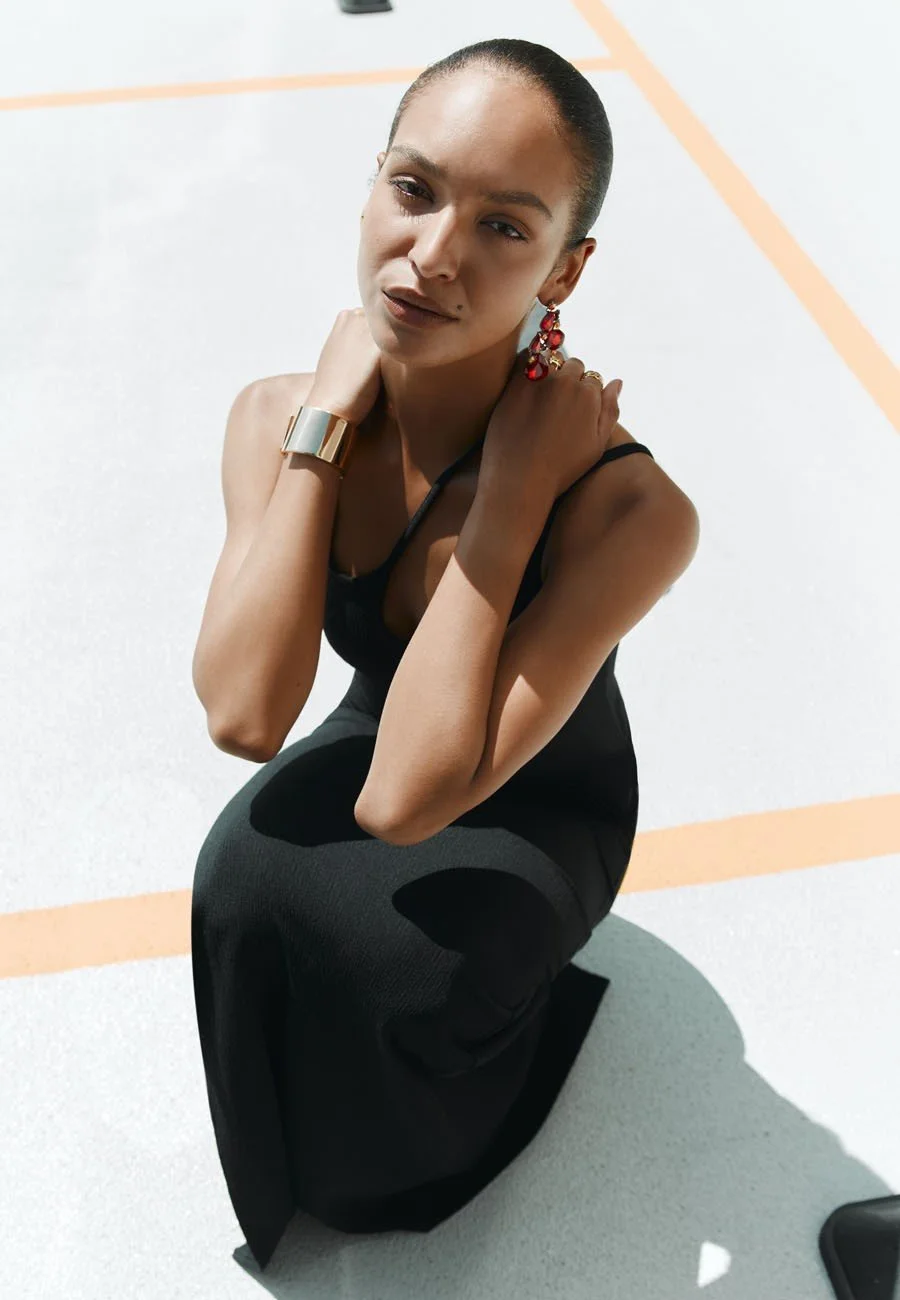A model kneeling in a car park, looking at the camera, in a black tank top and black pants, wearing red earrings and a metallic bracelet.