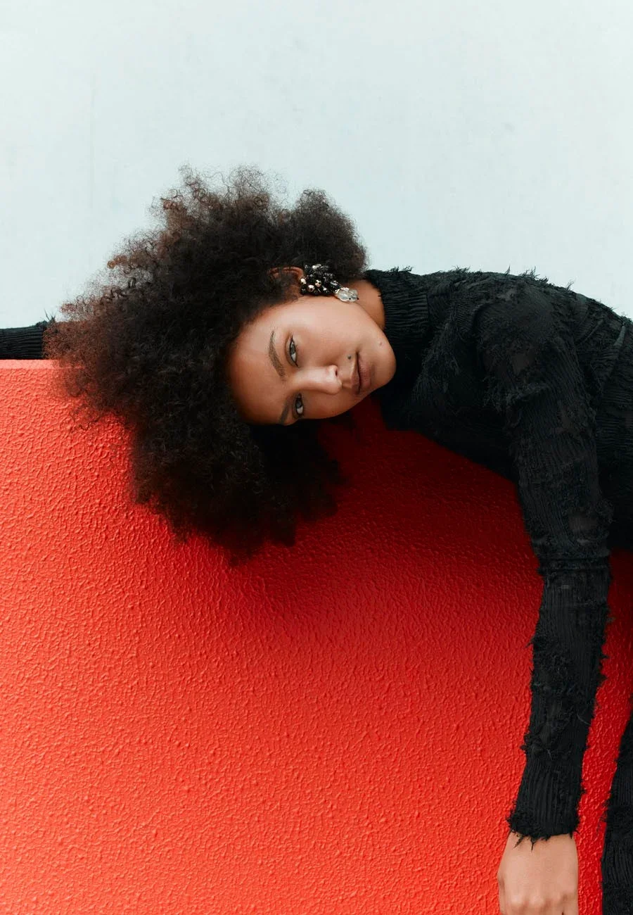 A model with curly hair lying sideways against a red textured wall, wearing a black top with distressed details, and earrings.