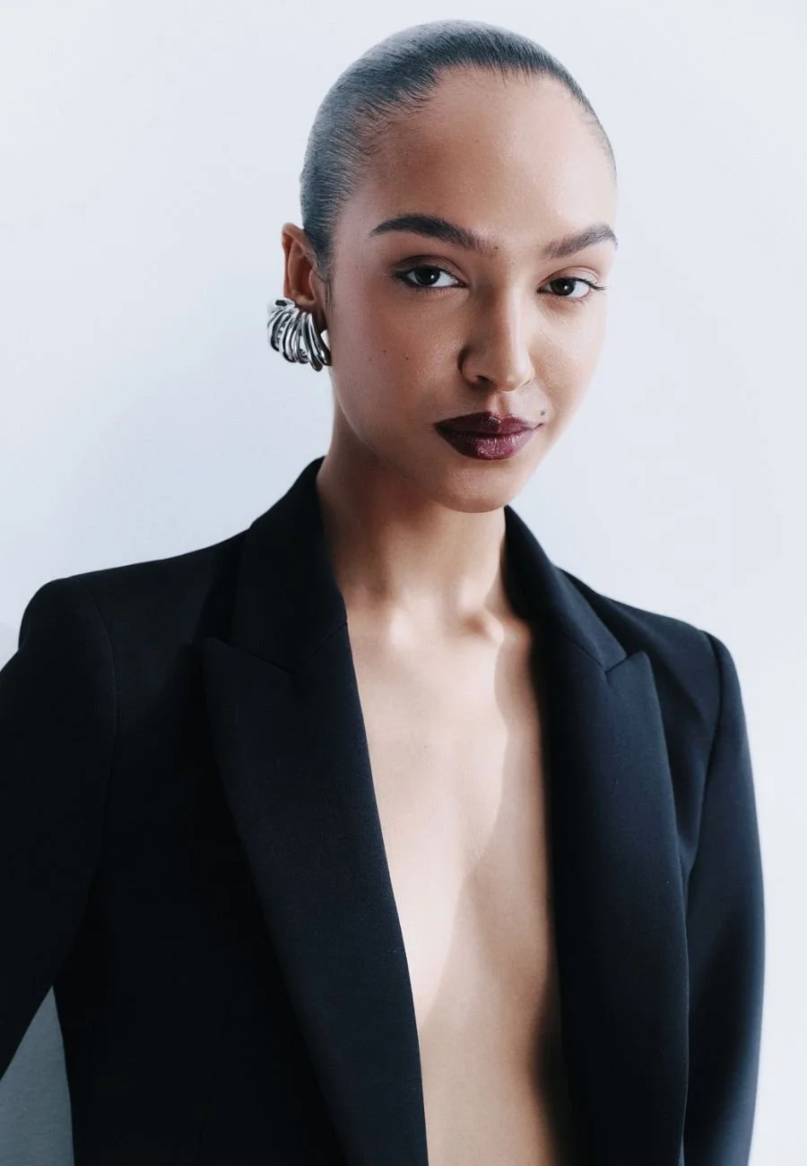 A model with her hair slicked back wearing a black blazer, dark lipstick, silver hoop earrings, and looking confidently at the camera.