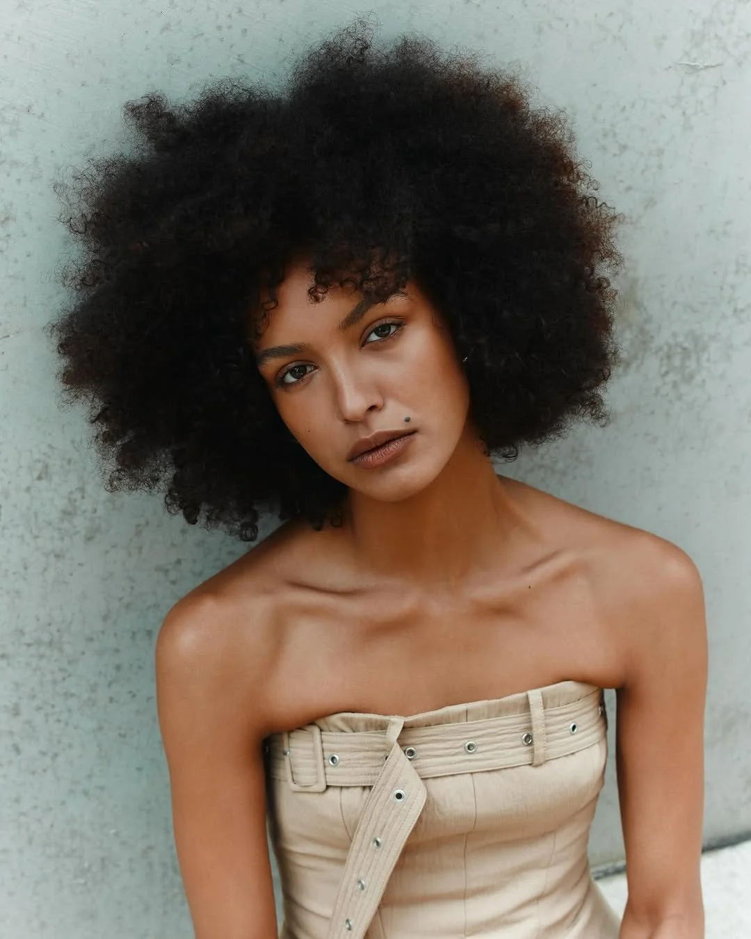 A model with curly natural hair and light brown skin leaning against a light-colored wall, wearing a beige strapless top.