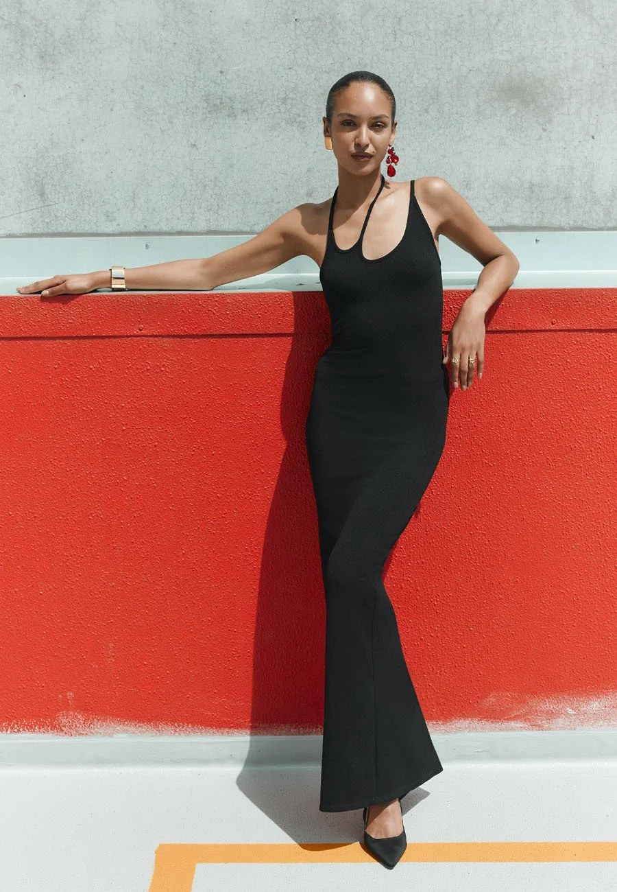 A model in a black dress and high heels standing against a red wall outside, with one arm resting on the wall and the other on her hip.