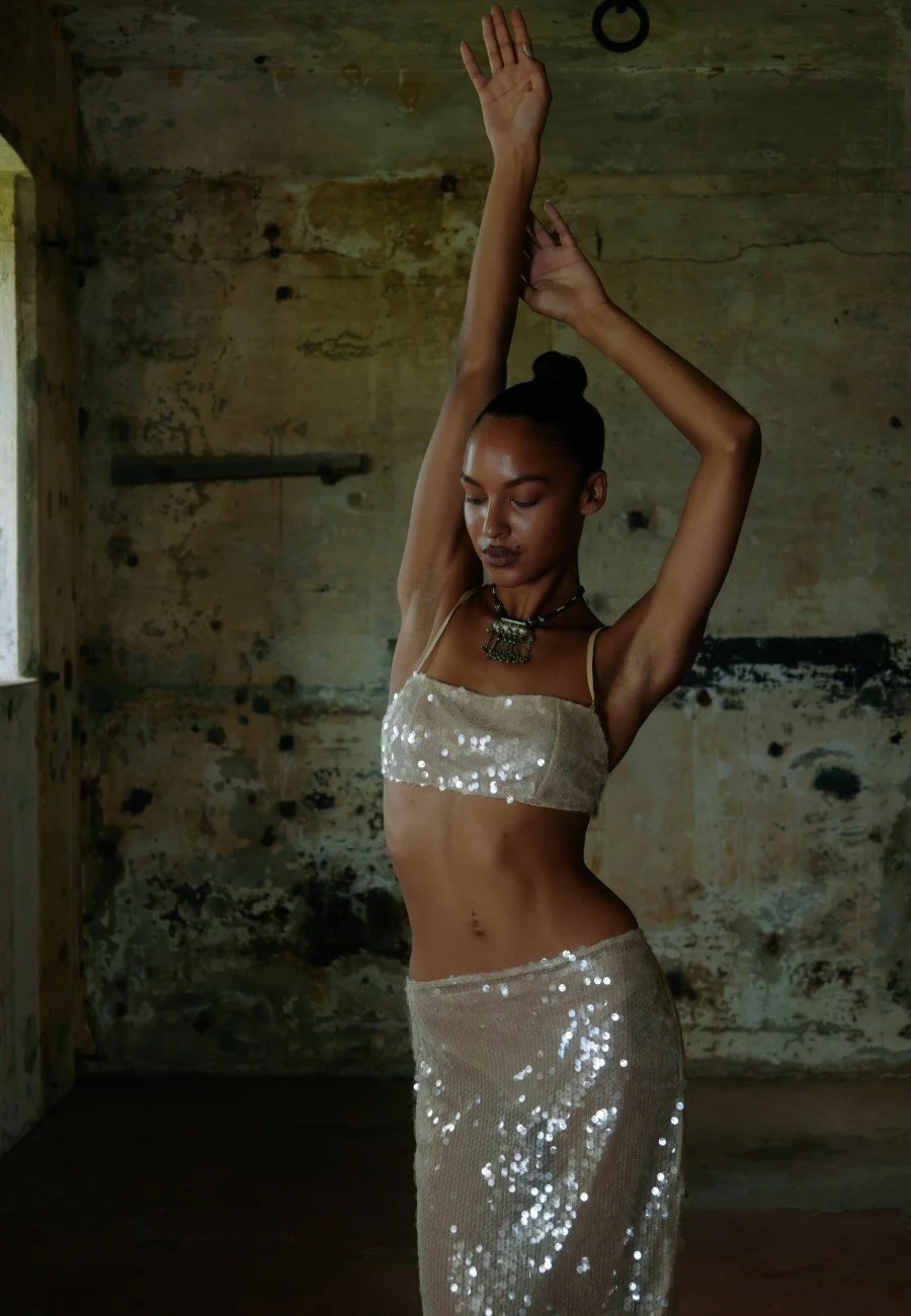 A woman with curly hair in a bun, wearing a shiny, sequined two-piece outfit, dances with arms raised inside a rustic, dimly lit room.