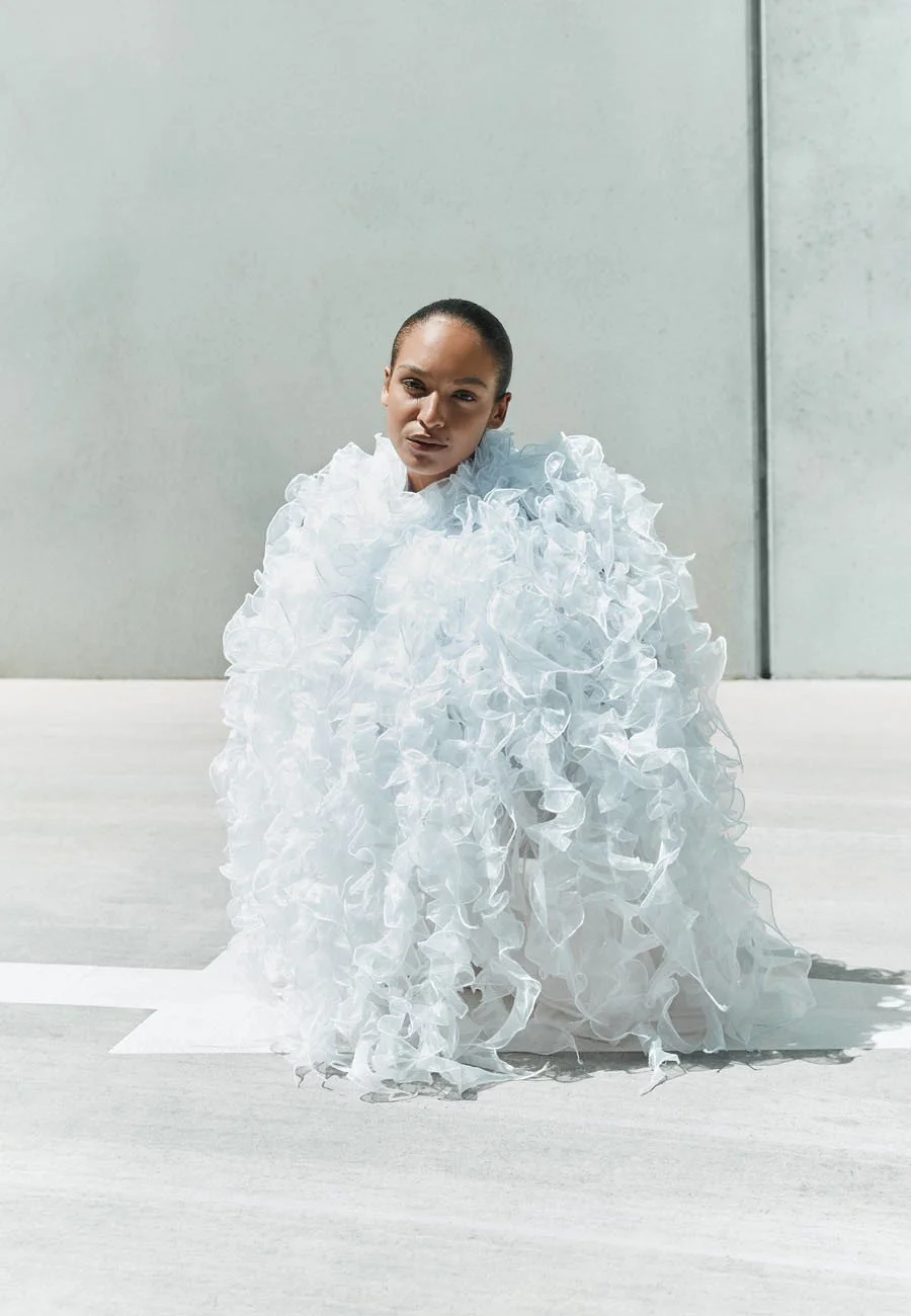 A model dressed in an elaborate white ruffled outfit, kneeling on a light-colored surface with a plain, textured wall behind her.