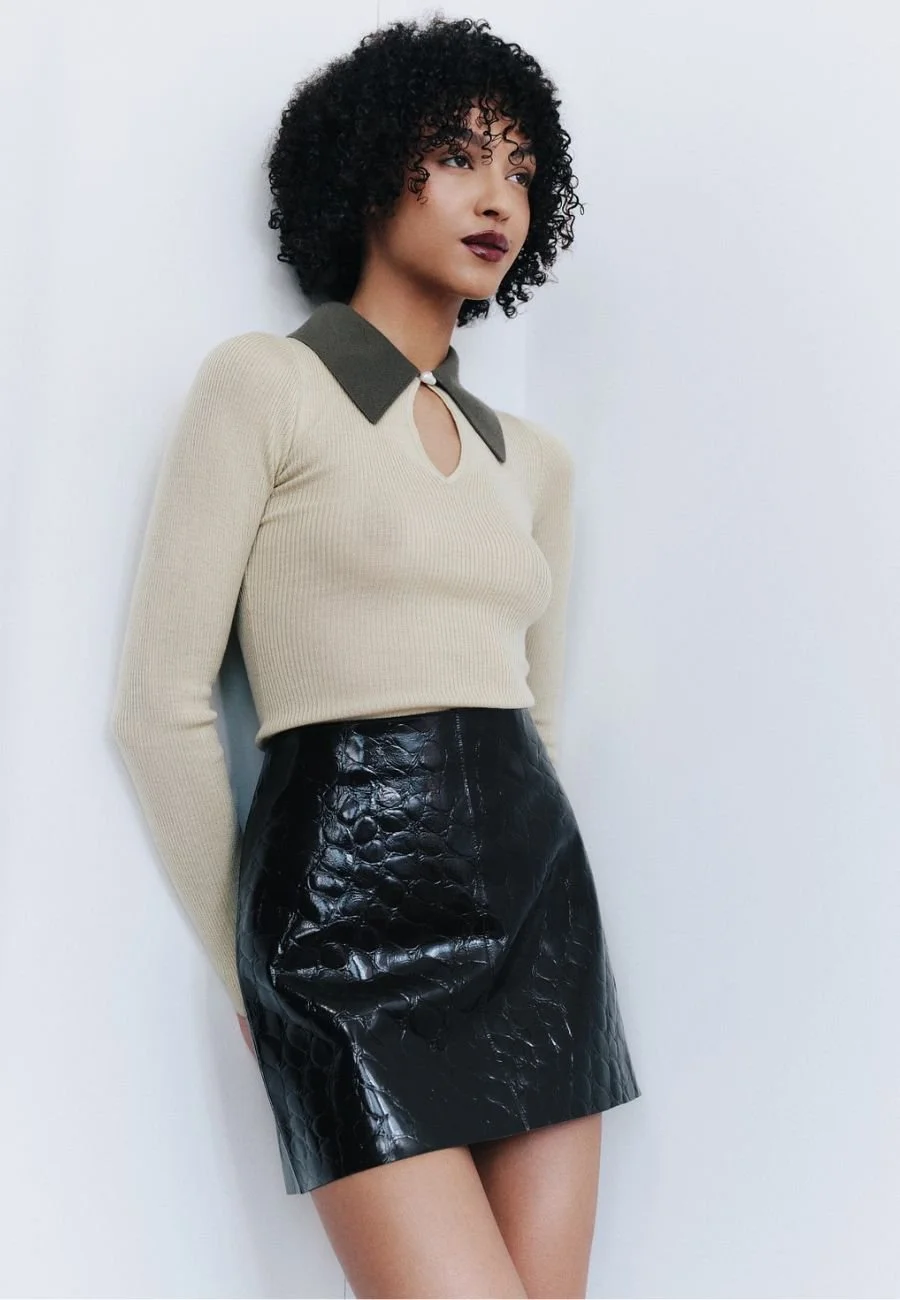 A woman with curly black hair wearing a beige long sleeve top with a dark collar and a keyhole cutout, paired with a black faux leather mini skirt, standing against a white wall.